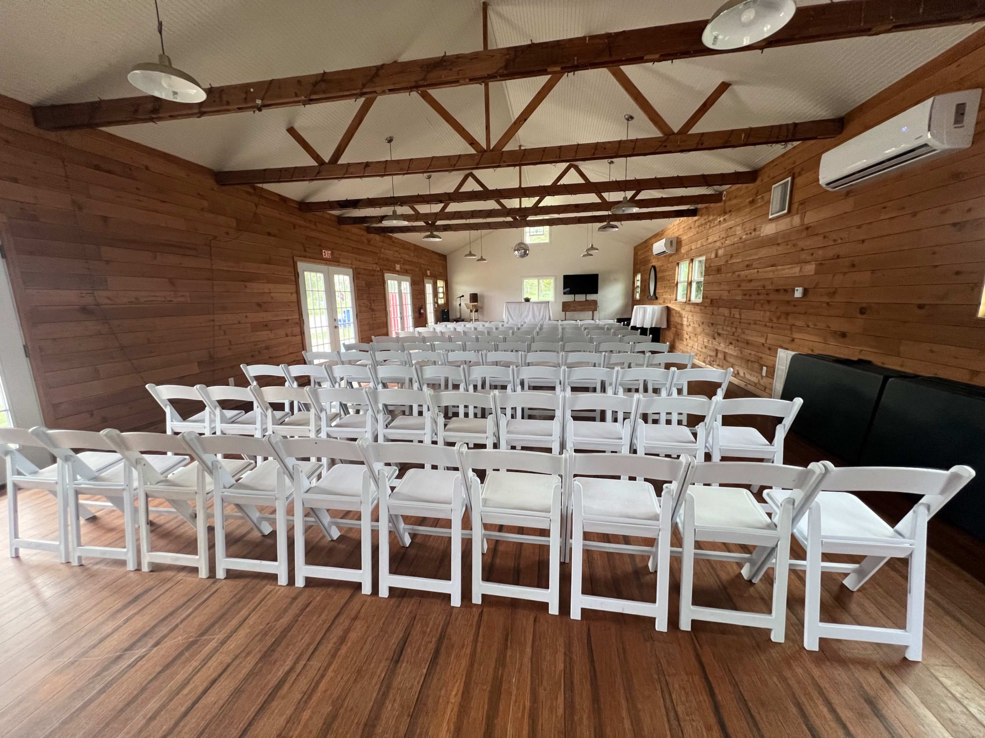 The image shows a spacious wooden room set up with rows of white folding chairs arranged for an event.