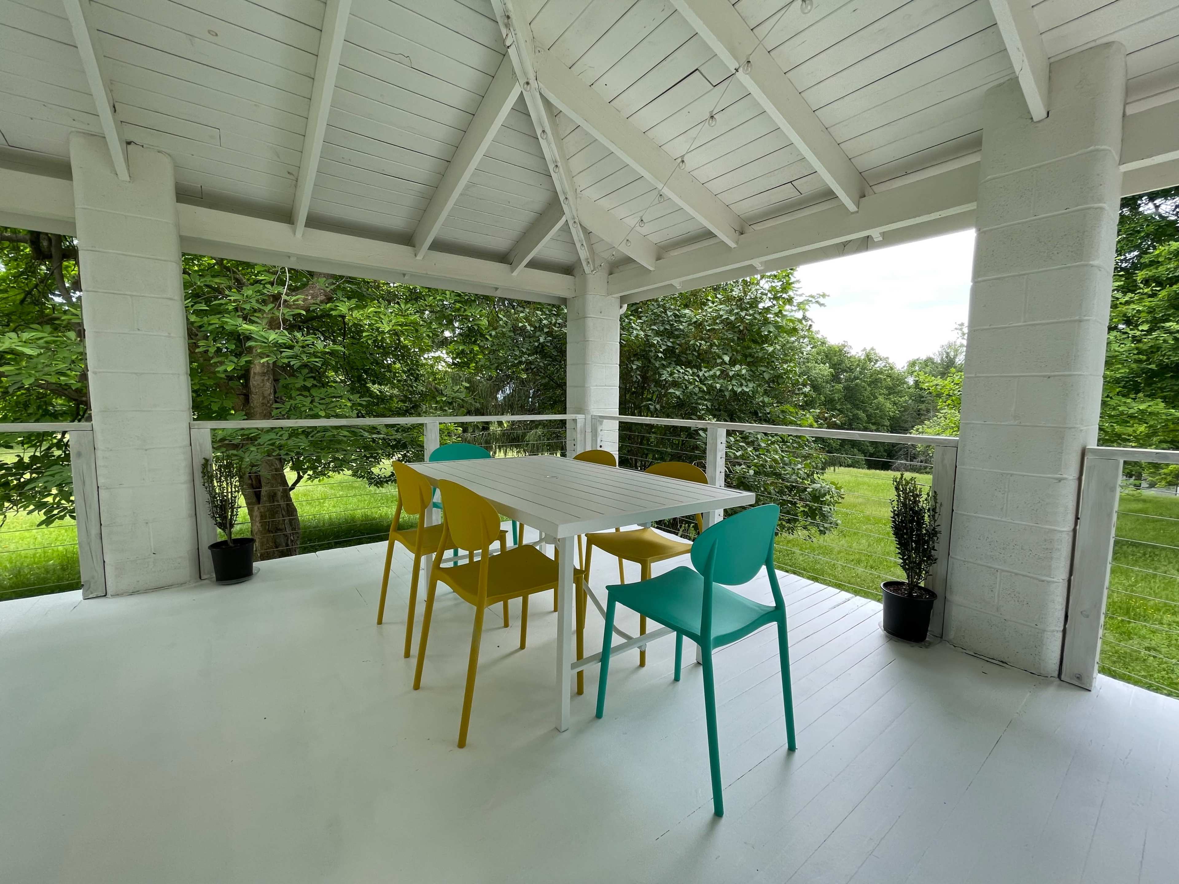 A bright, open gazebo features a white table surrounded by six brightly colored chairs overlooking a grassy area.