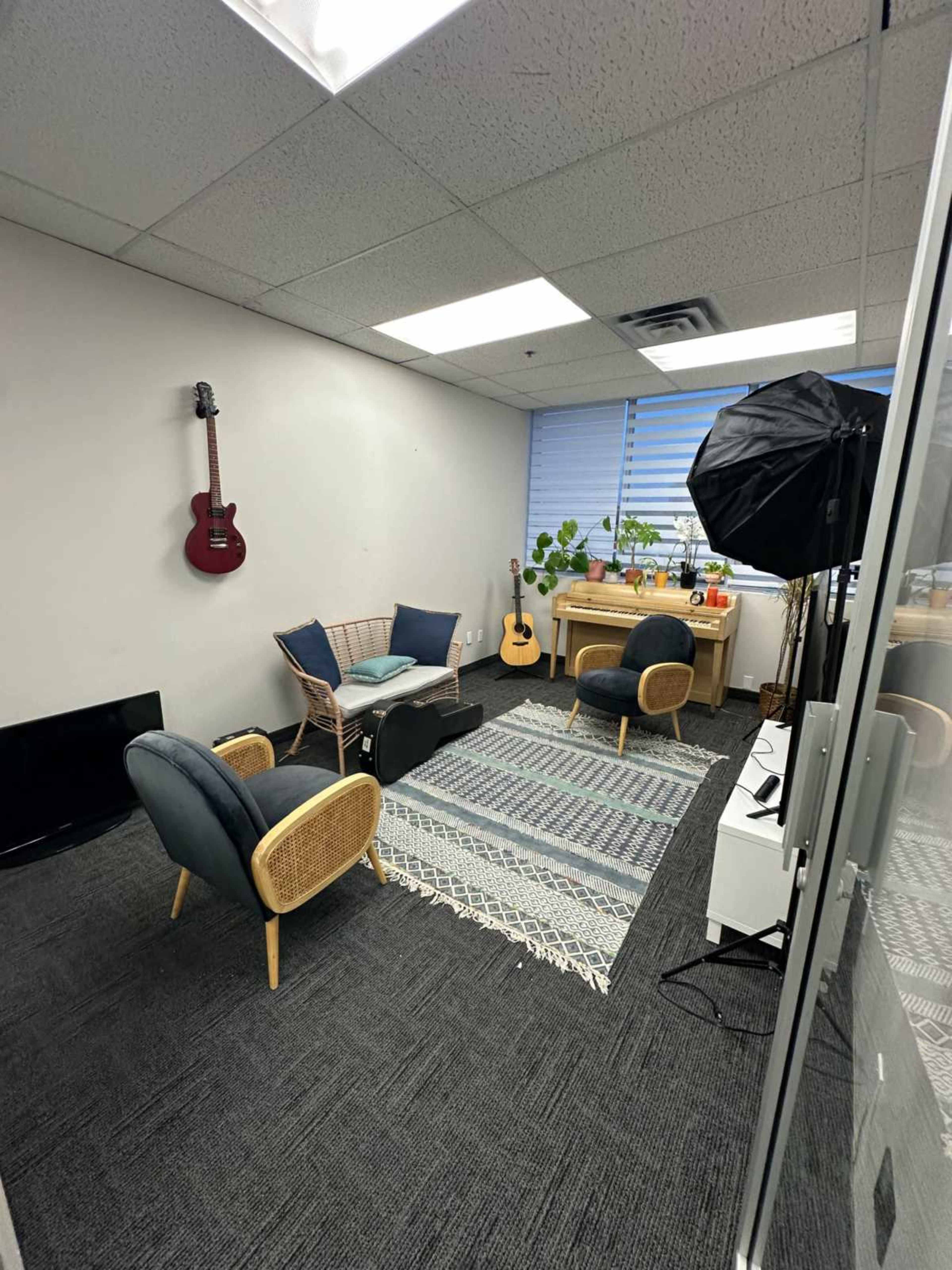 The image shows a small, cozy room with two chairs, a couch, a rug, plants, musical instruments, and a desk.