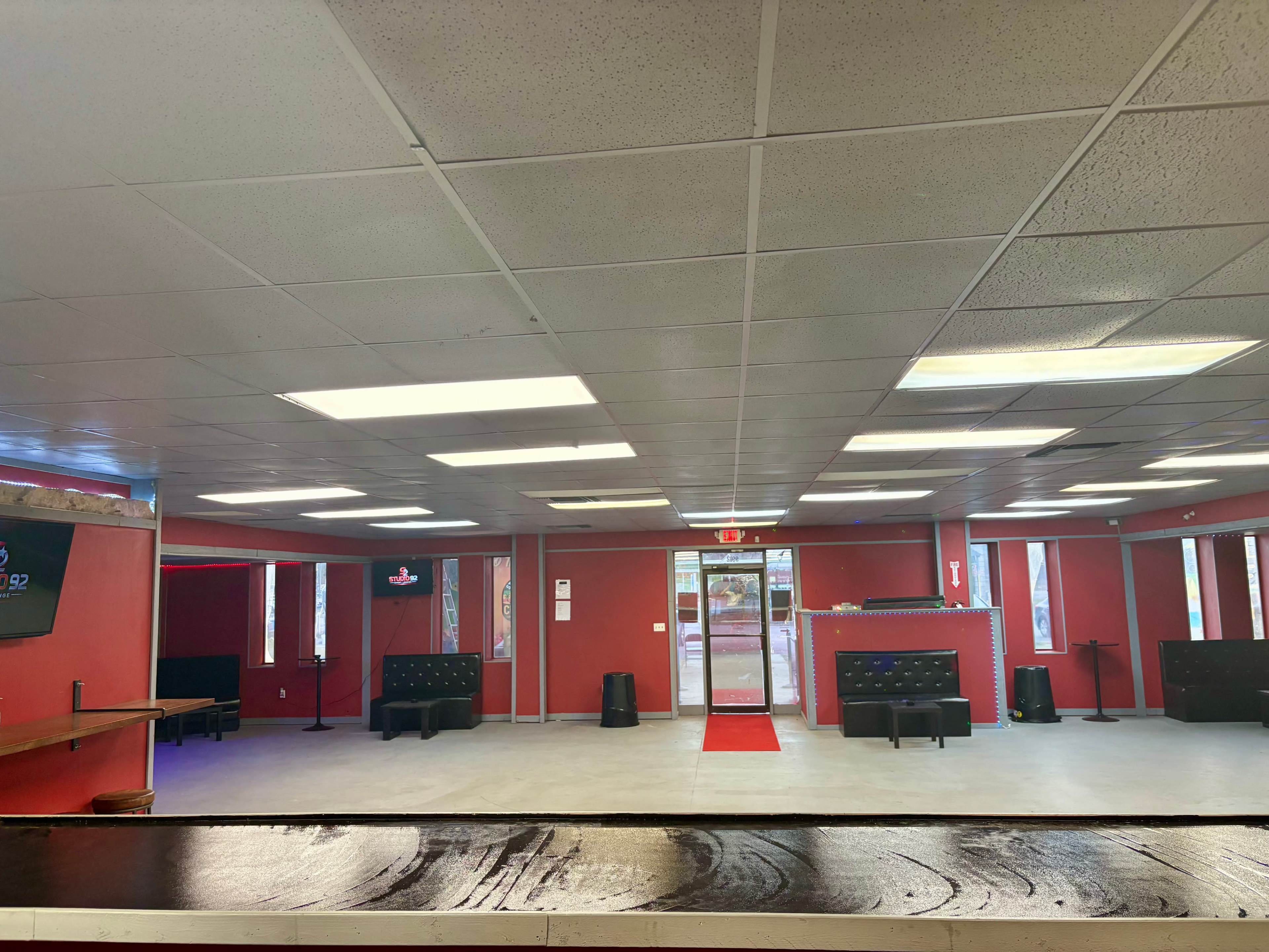 The image shows a modern interior of a lounge-like space with red walls, black seating, and a prominent red carpet leading towards an entrance.