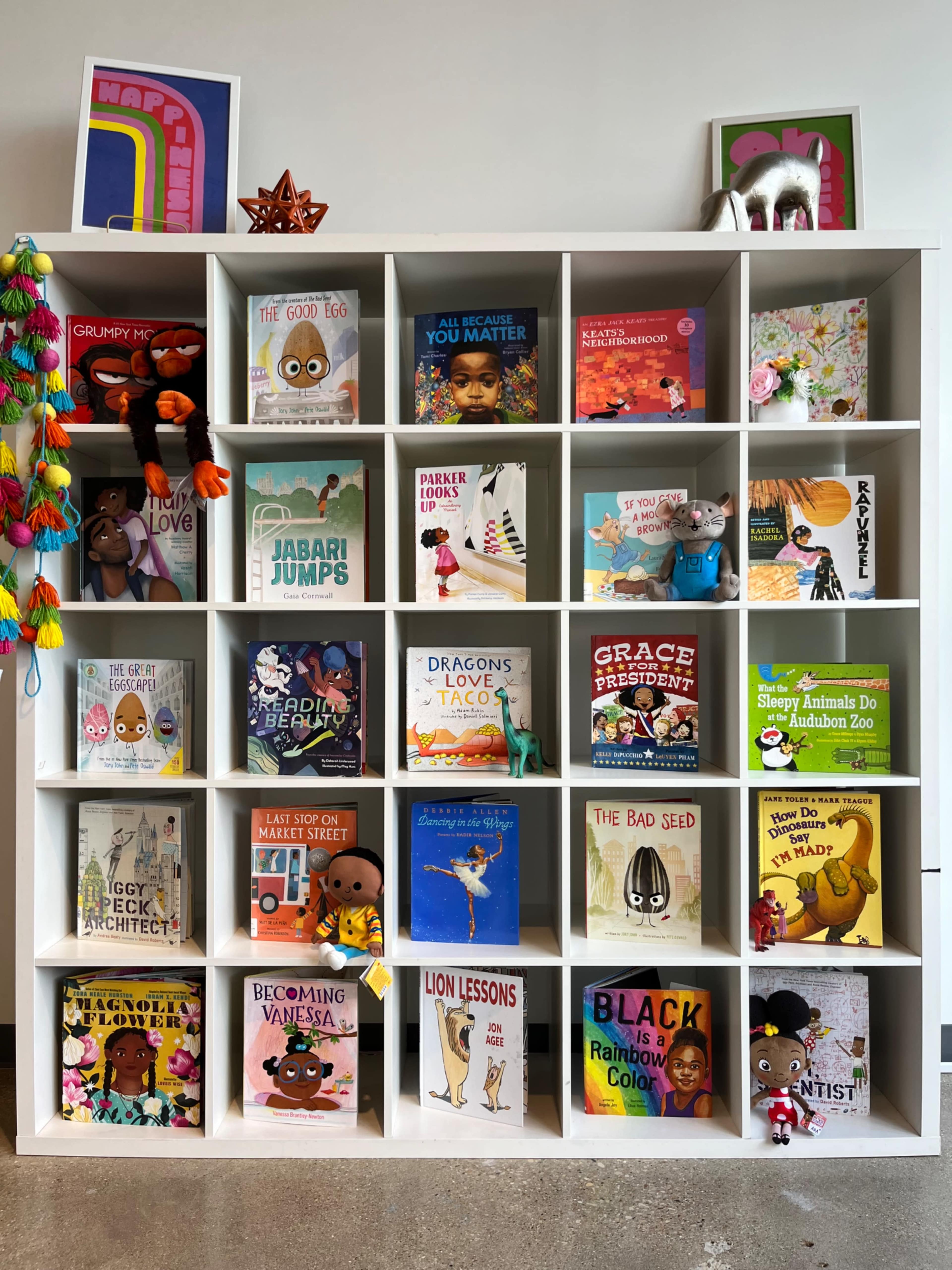 A white shelf displays a variety of children's books, organized in a grid with colorful covers and decorative items.