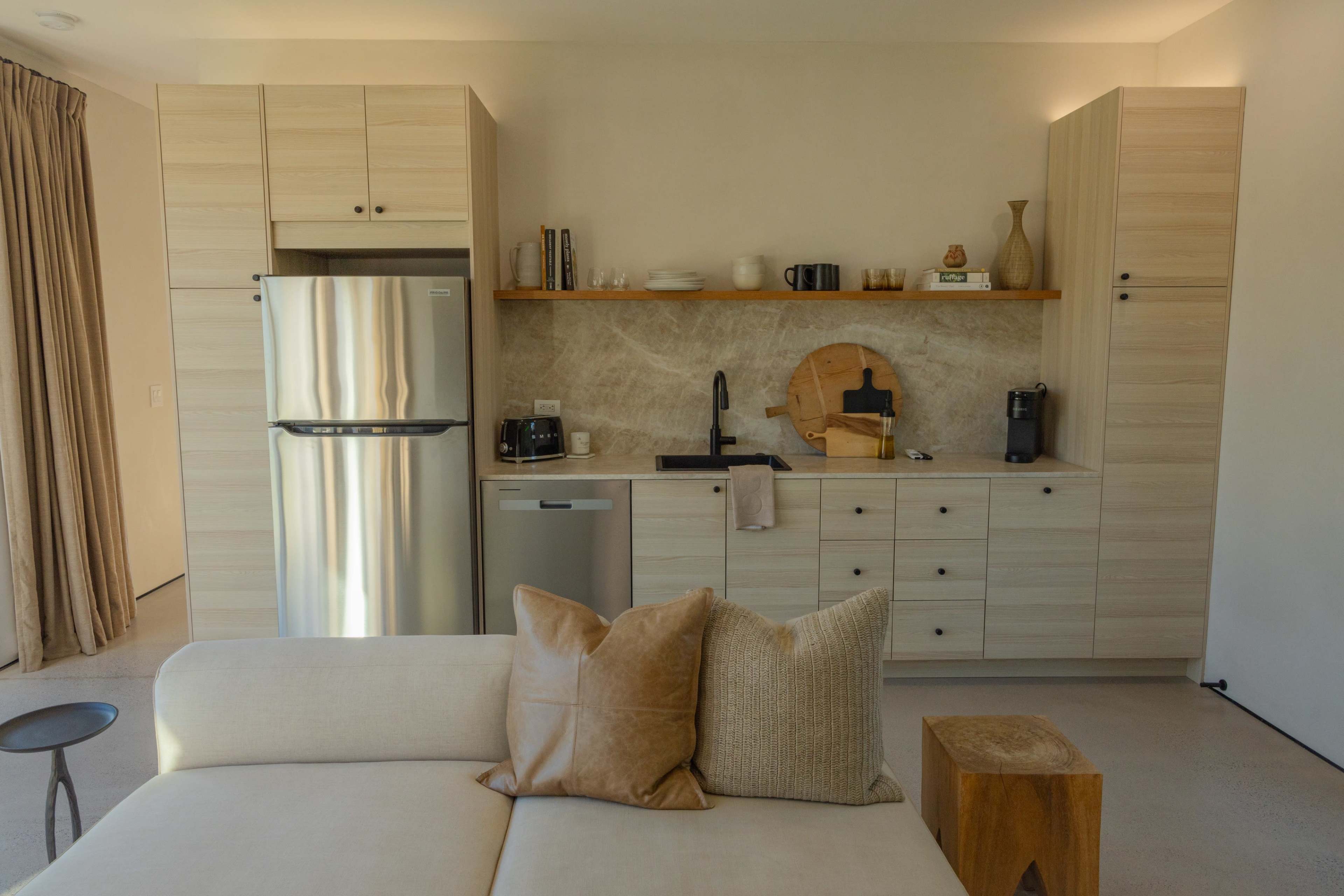 A modern kitchen features light wood cabinetry, a stainless steel refrigerator, a sink, and a small dining area with a sofa in the foreground.