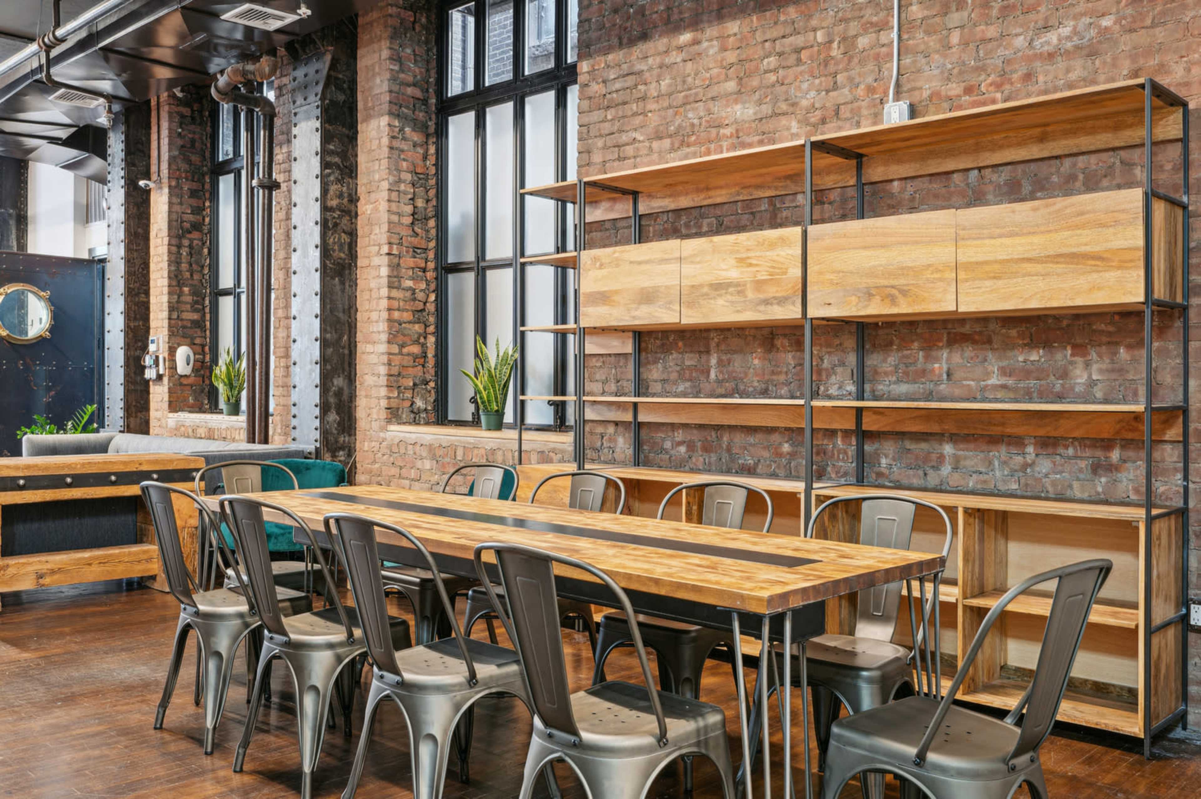 The image shows a modern dining area with a long wooden table surrounded by metal chairs, set against a backdrop of exposed brick walls and large windows.