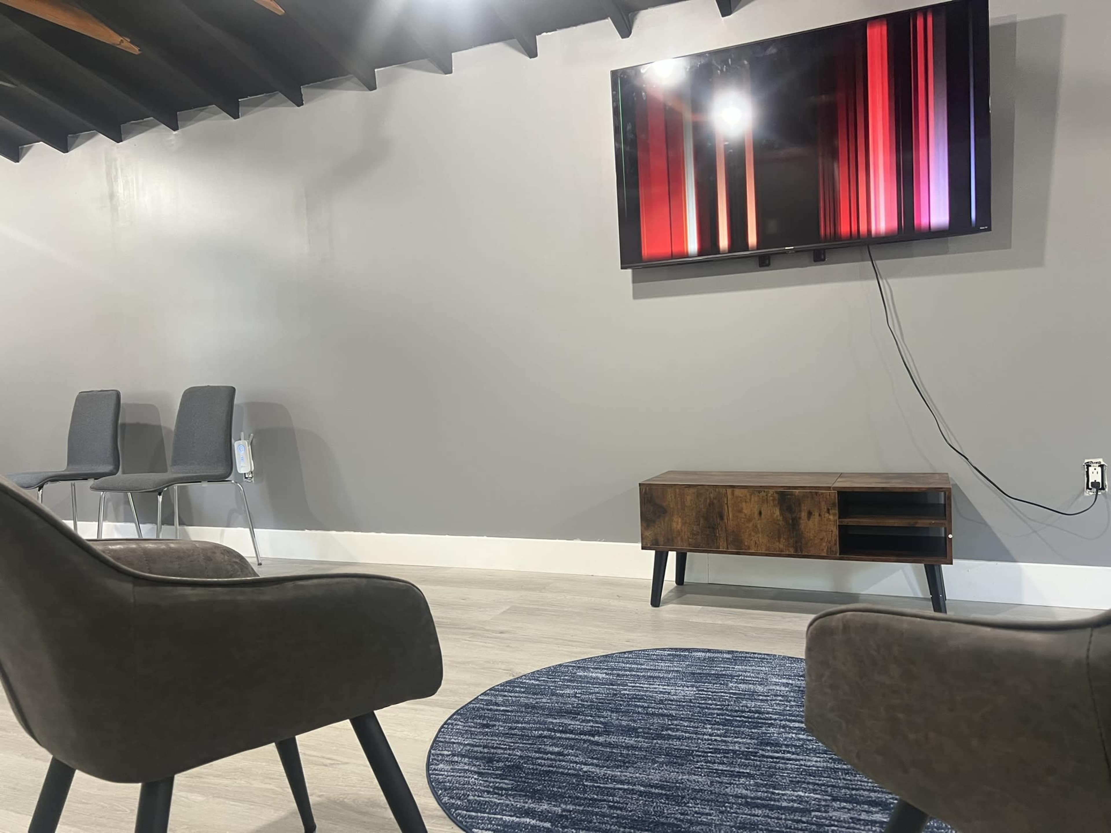 The image shows a modern waiting area featuring two chairs facing a wall-mounted television displaying colorful vertical stripes, with a wooden cabinet below the TV and a circular blue rug on the floor.