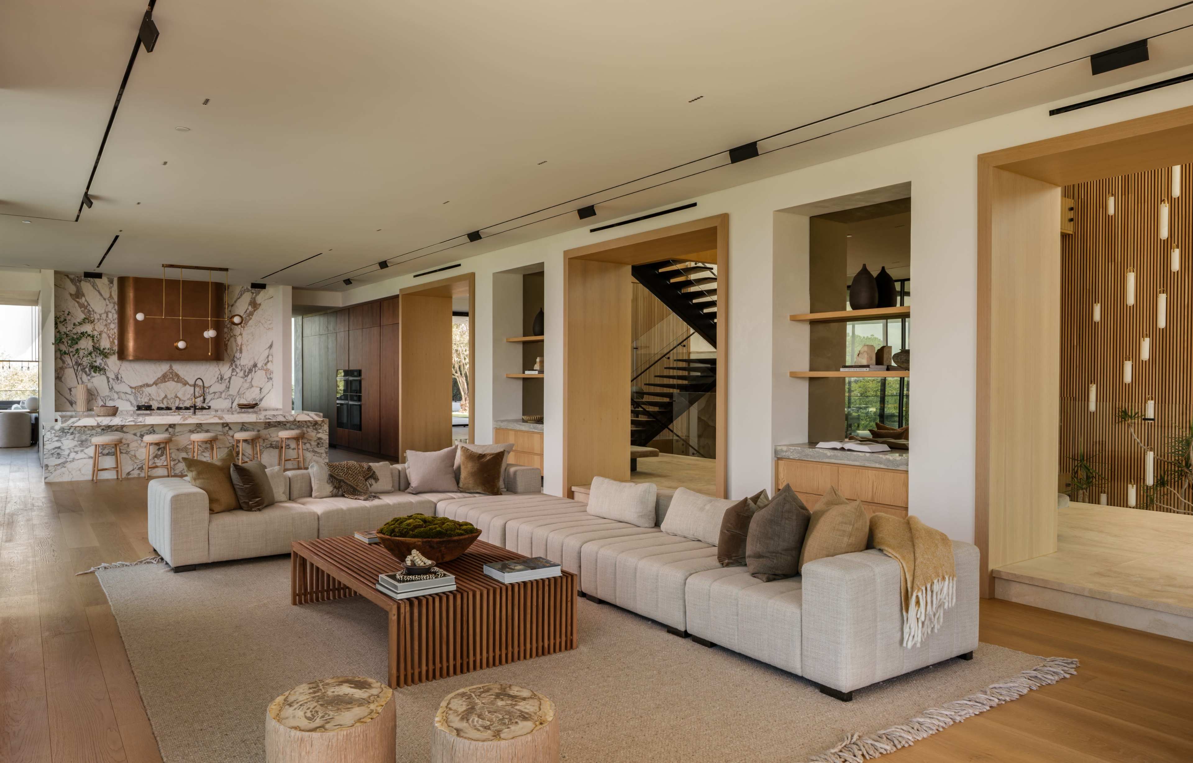 The image shows a spacious, modern living room featuring a large sectional sofa, a wooden coffee table, and an open kitchen area with a marble backsplash.