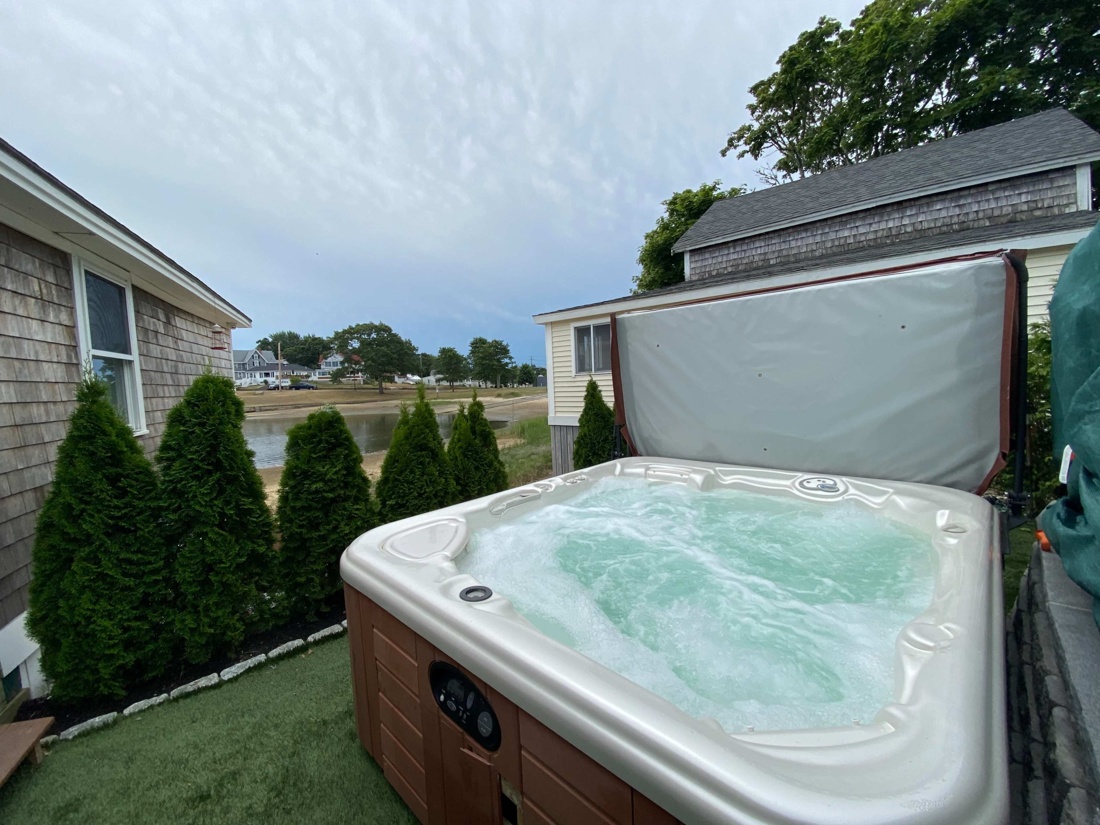 A hot tub filled with water is positioned on a patio surrounded by tall green shrubs, with a view of a lake in the background.