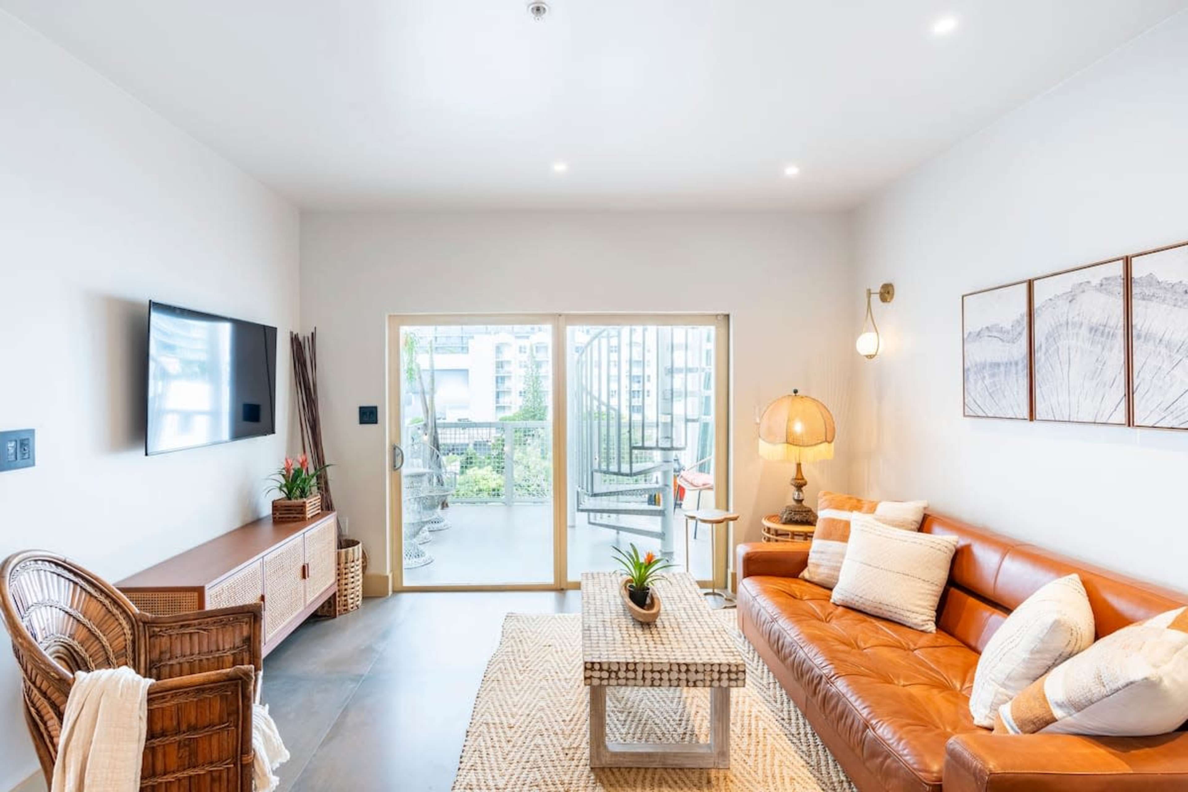 The image shows a modern living room with a brown leather sofa, a wooden entertainment unit, and large glass doors leading to a balcony.