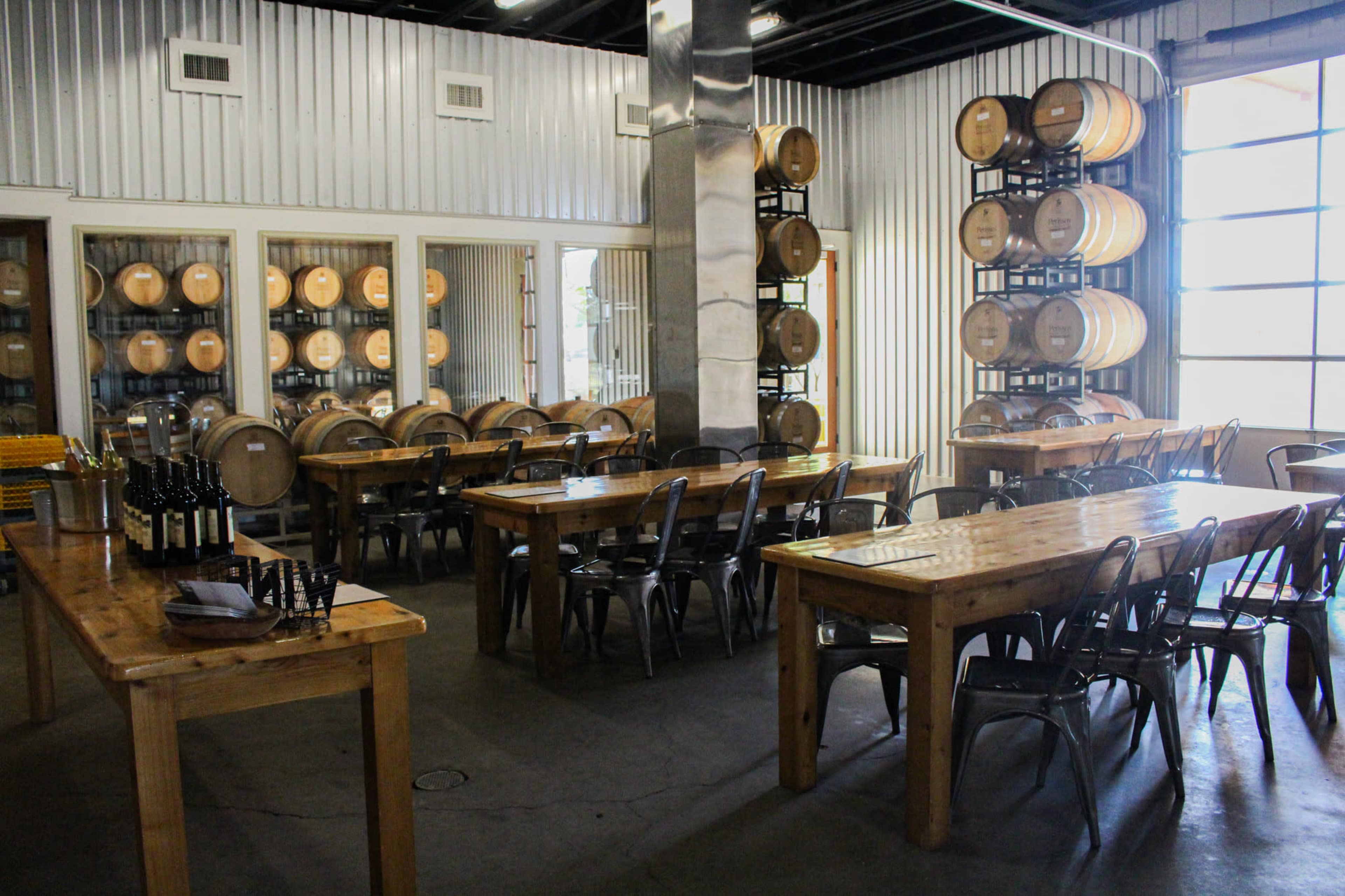 A spacious winery tasting room with wooden tables, metal chairs, and wine barrels stacked in the corner.