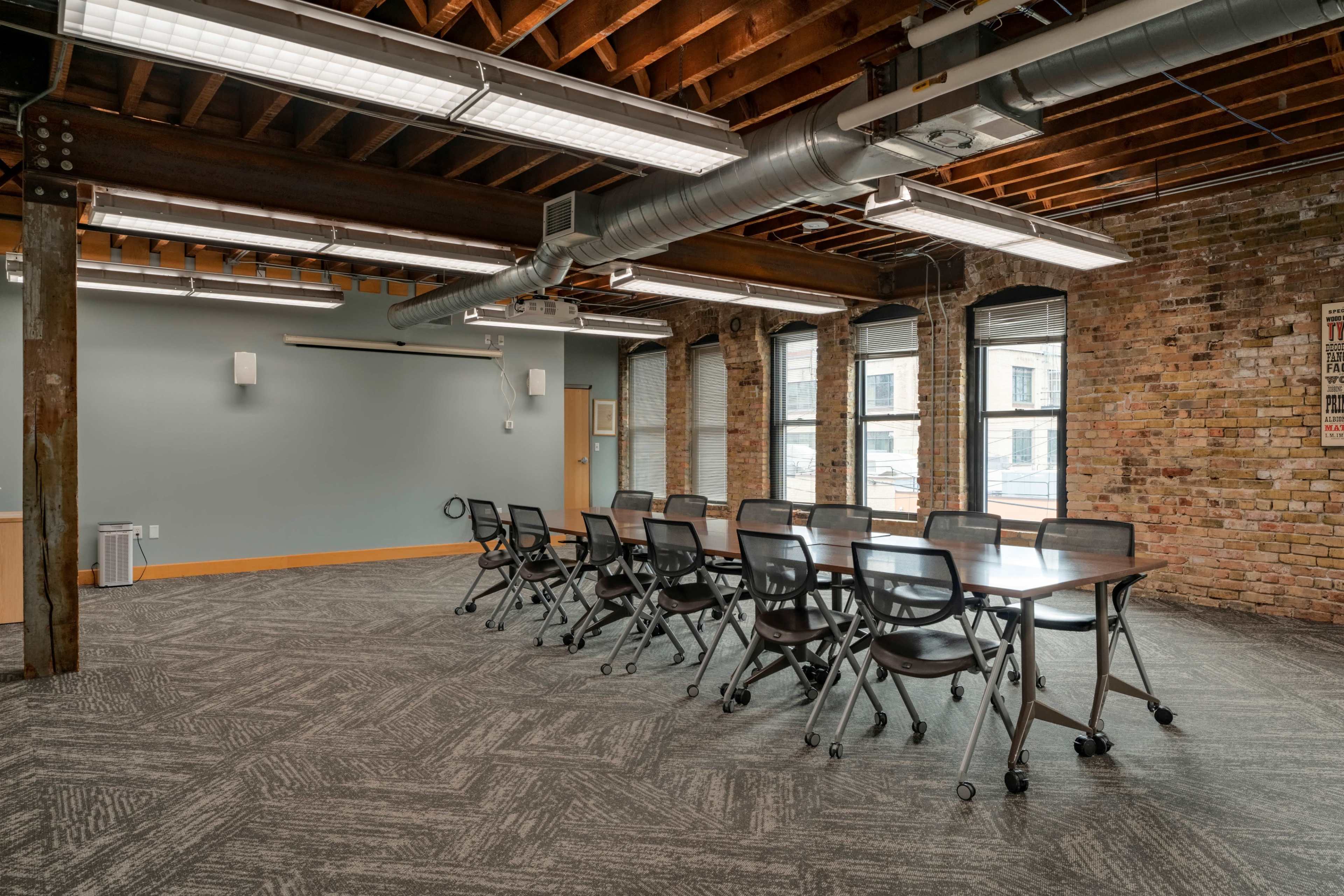A modern conference room with a long table surrounded by black rolling chairs, set against brick walls and exposed wooden beams.