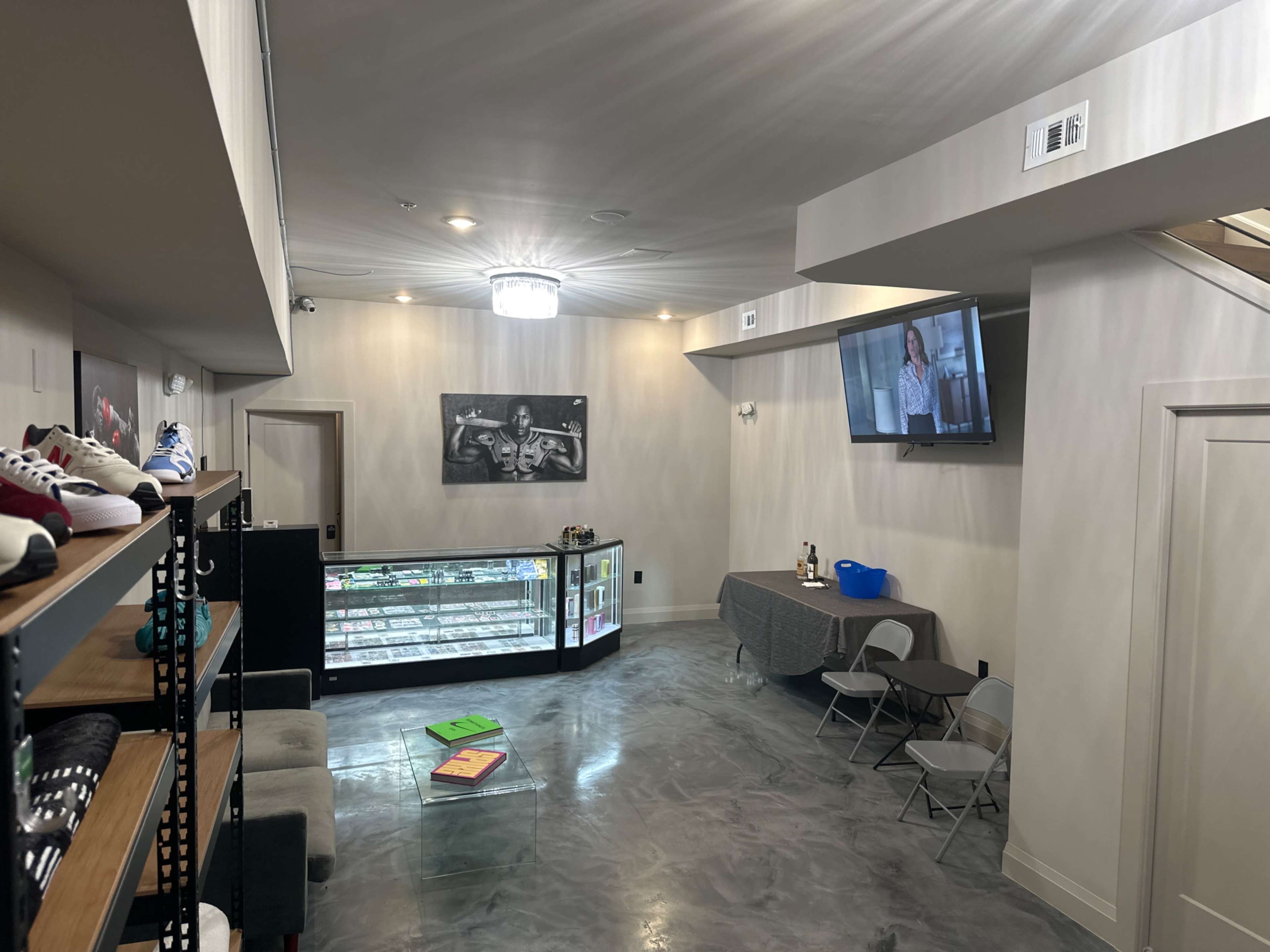 A modern retail space with concrete flooring, a display rack of shoes on the left, a table with chairs in the center, and a wall-mounted television in the corner.
