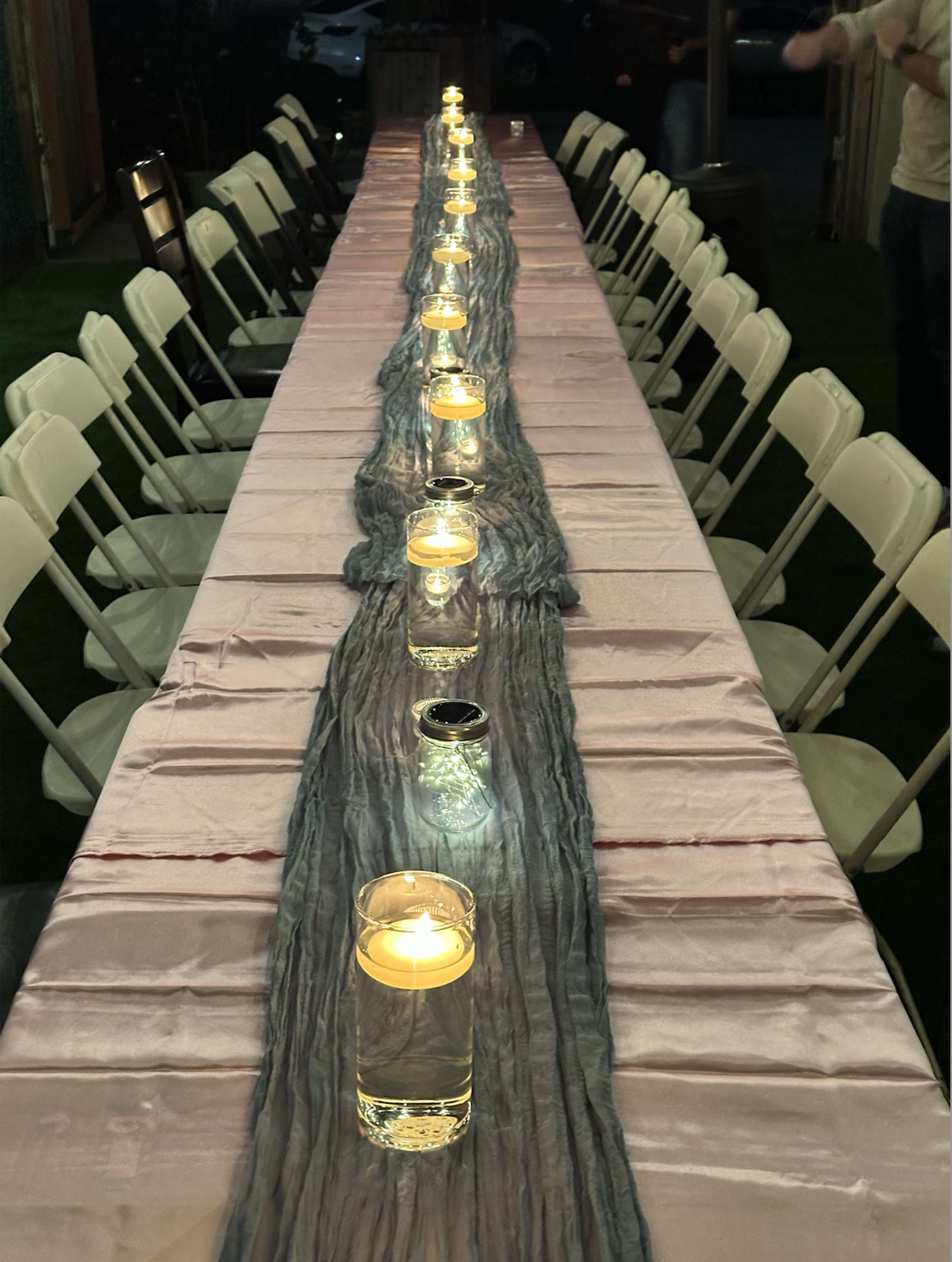 A long table is set outdoors with a light pink tablecloth, flanked by white chairs and illuminated by candles in glass holders.