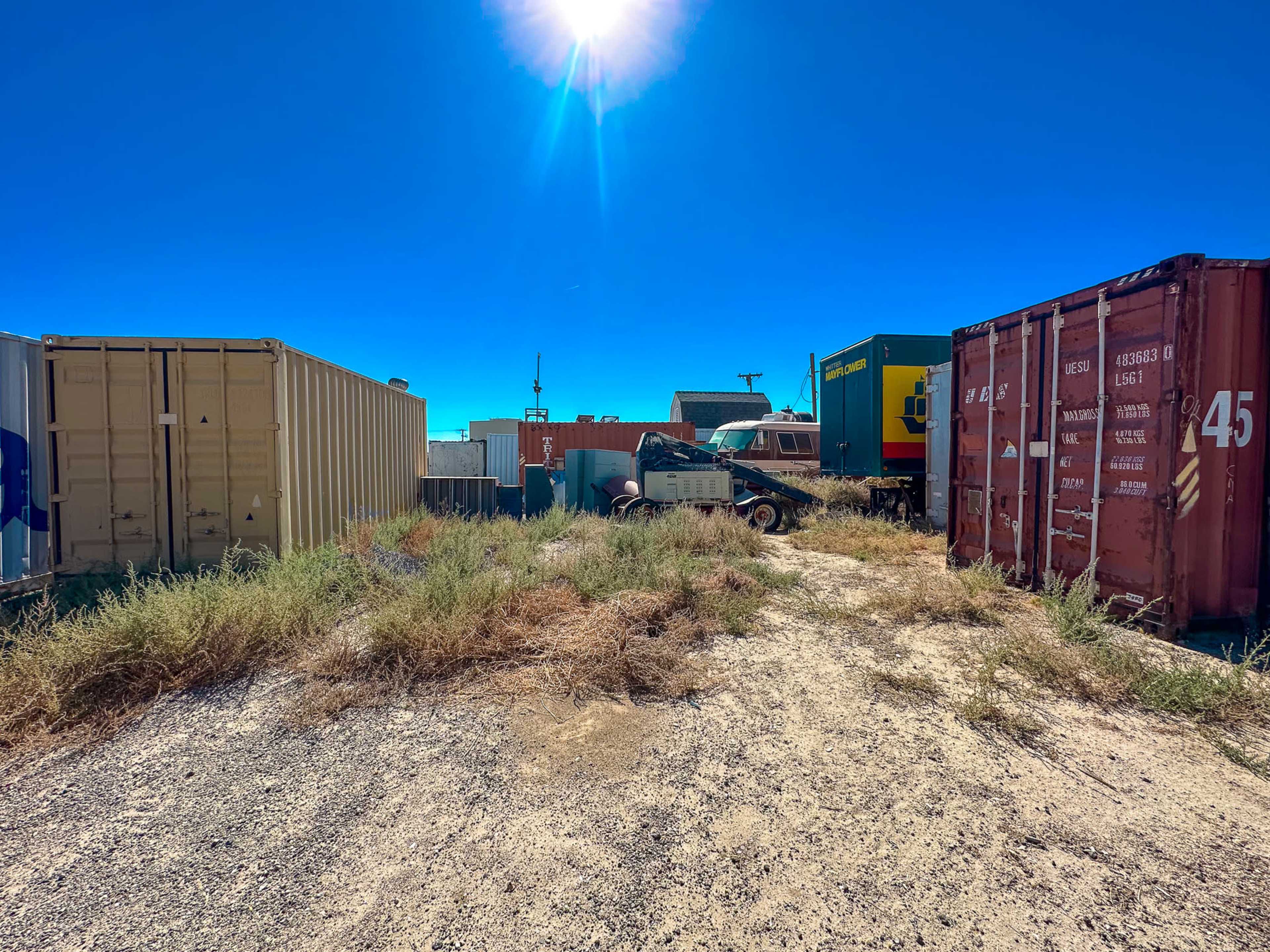 The image shows a collection of colorful shipping containers surrounded by dry grass and gravel under a clear blue sky.