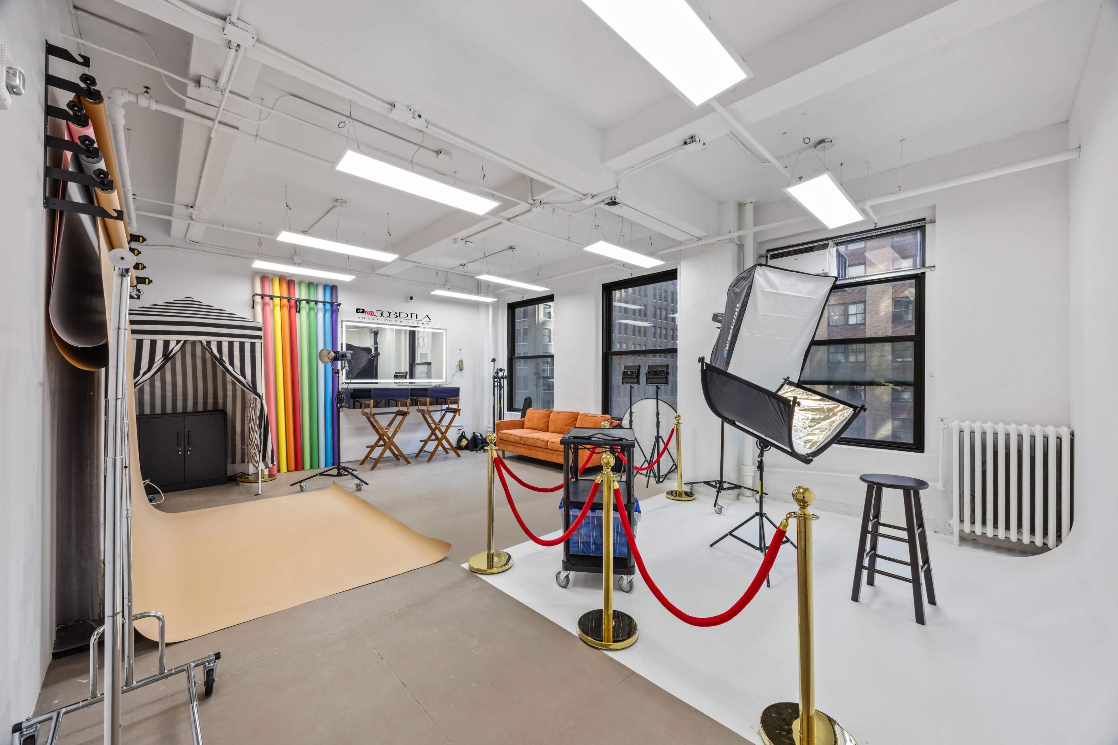 The image shows a brightly lit photography studio featuring a backdrop area with colorful rolls, a reception area with an orange couch, and various lighting equipment set up.