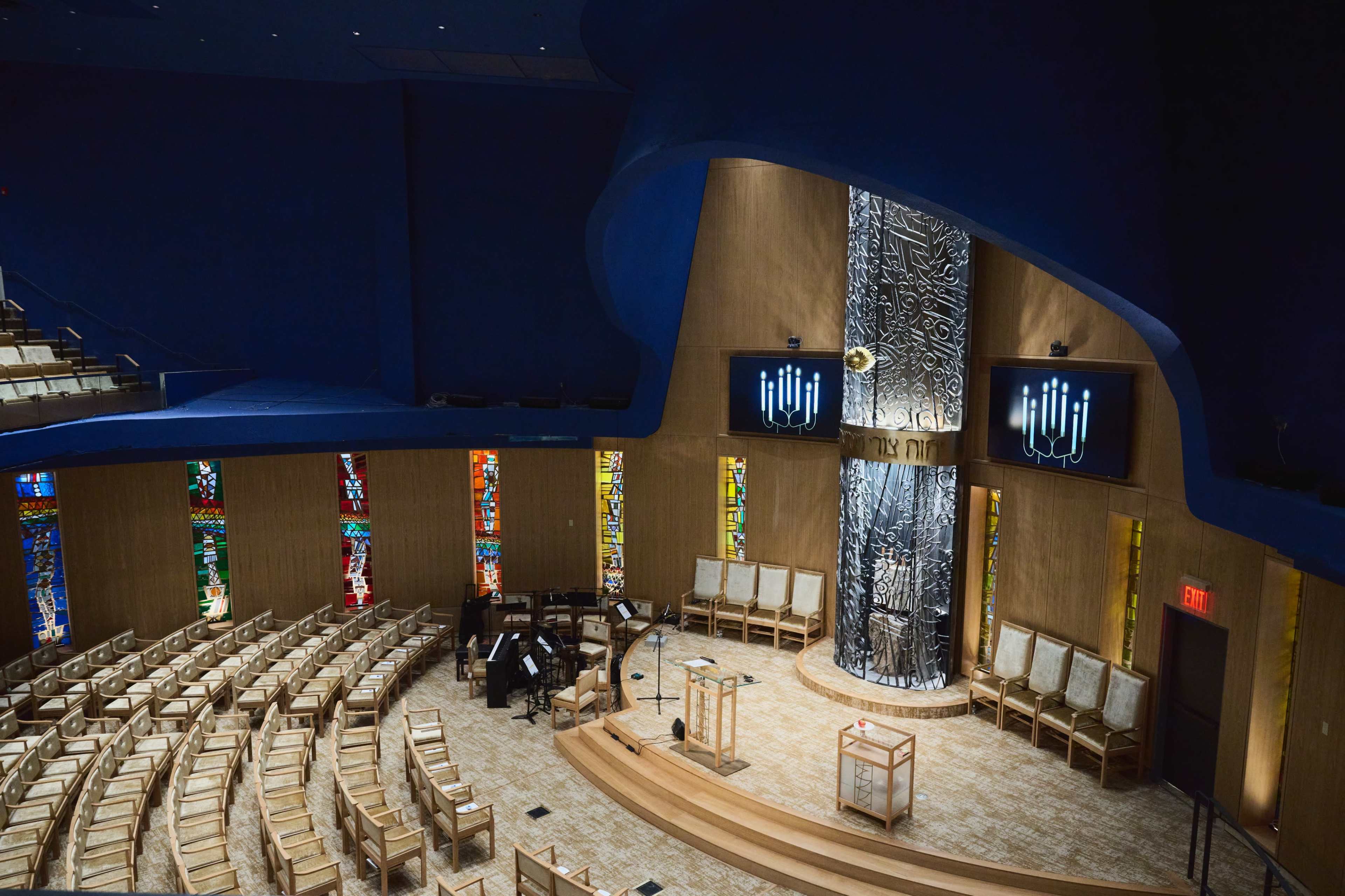 The interior of a modern synagogue featuring colorful stained glass windows, a central wooden platform, and large screens displaying inscriptions.