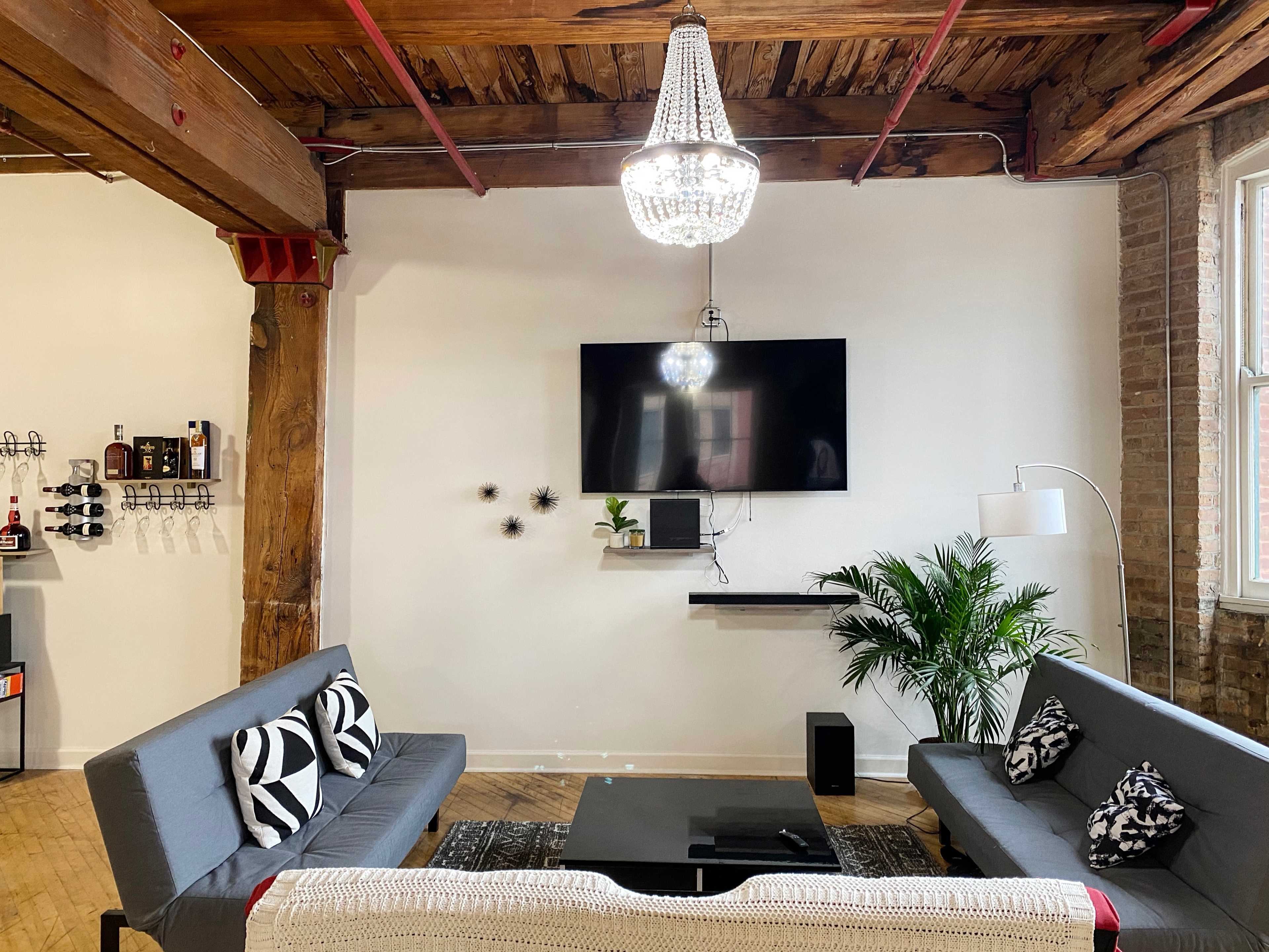 A modern living area features two gray sofas, a black coffee table, a flat-screen TV mounted on the wall, and ornamental plants.