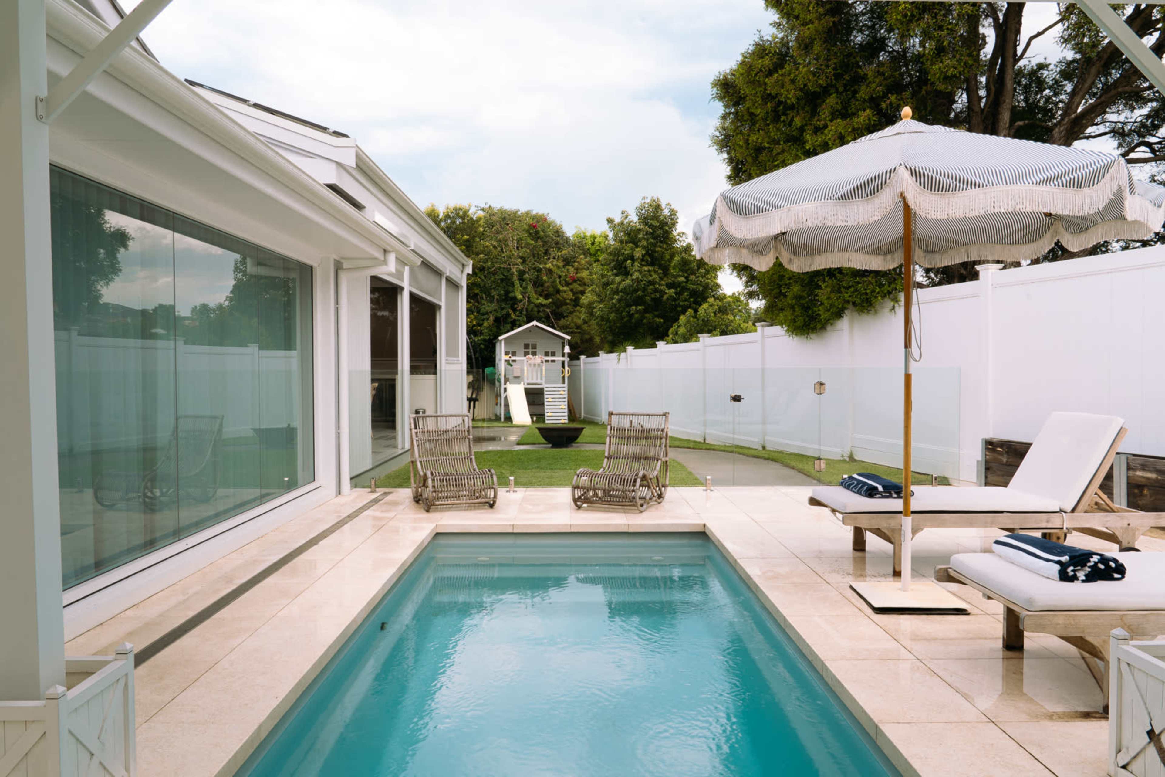 The image features a modern patio with a small swimming pool, surrounded by lounge chairs and a large umbrella, overlooking a well-maintained lawn with a playhouse in the background.