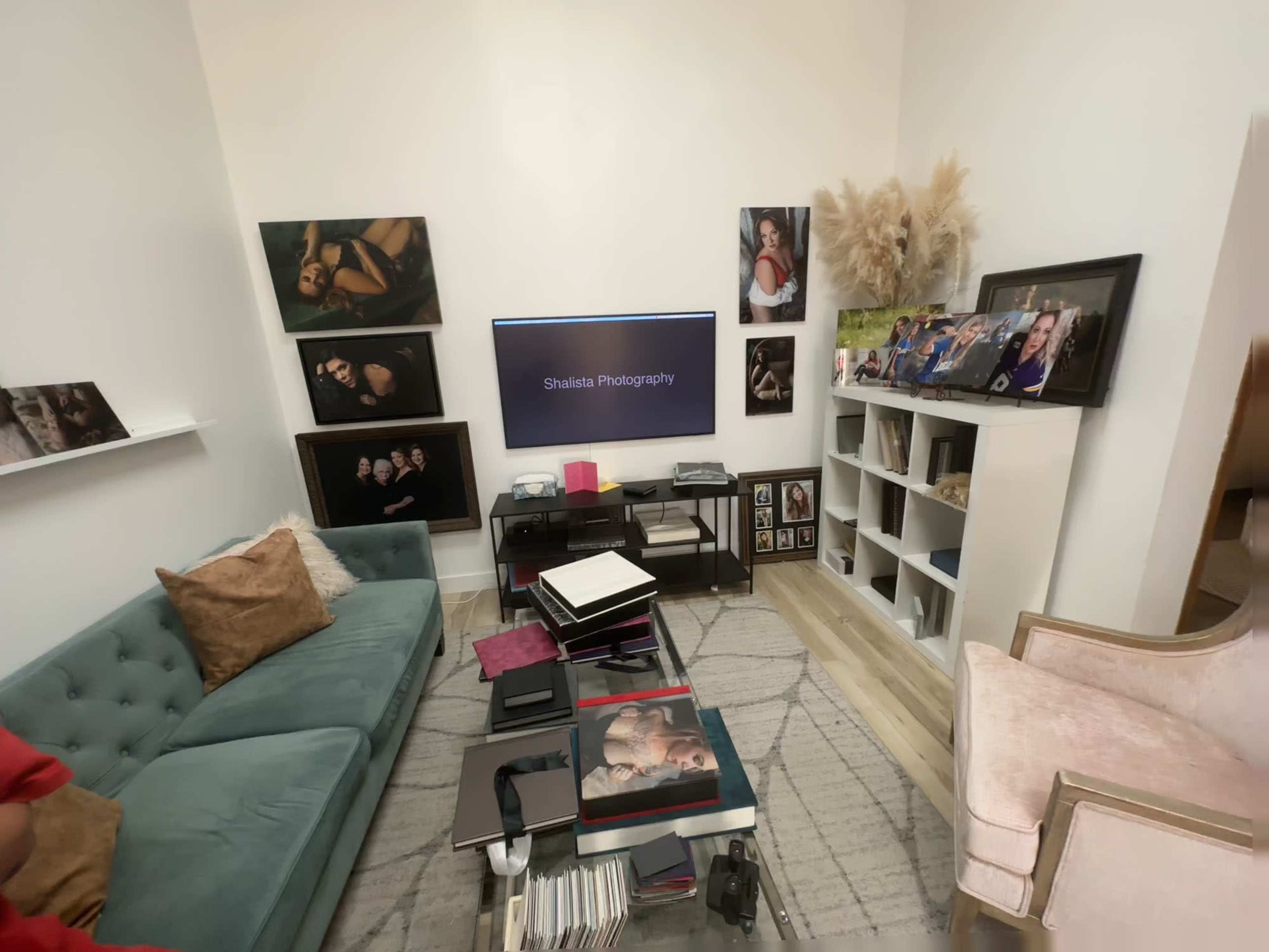 A cozy photography studio with a green couch, a television displaying "Shakira Photography," various framed photographs on the walls, and a bookshelf filled with photo albums.