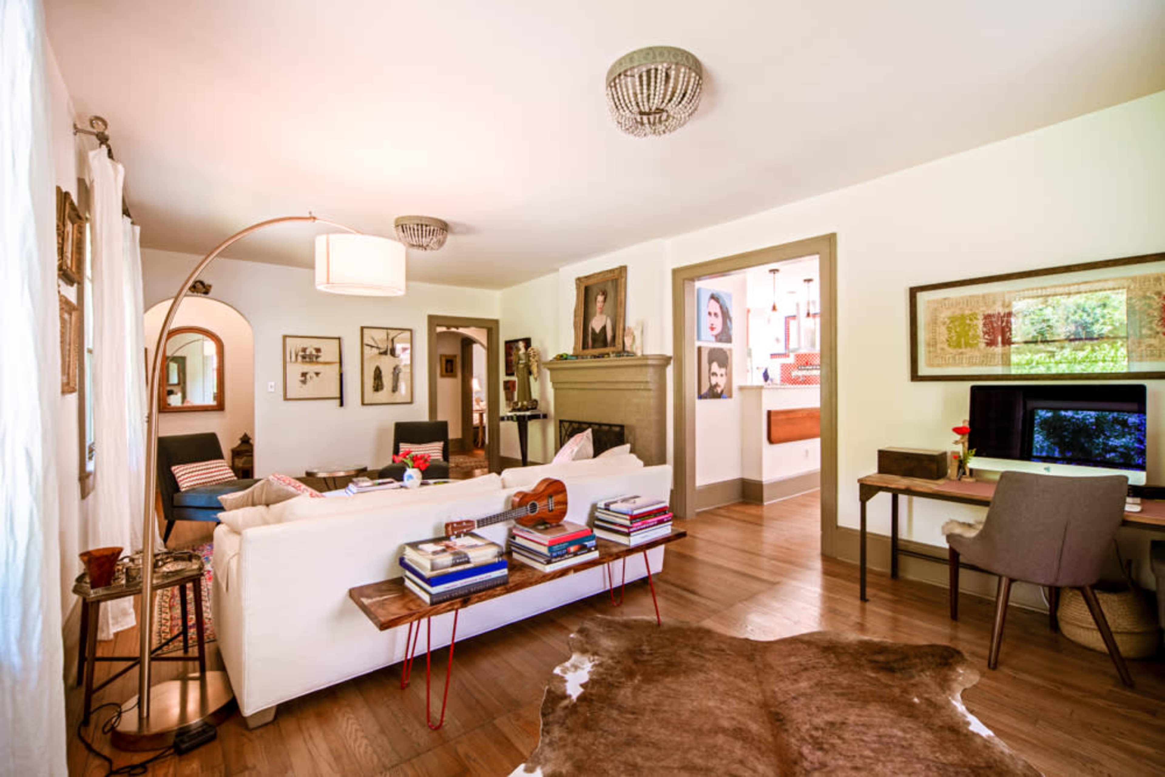 The image shows a cozy living room with a white sofa, wooden floors, and a desk, featuring various decorative elements and framed artwork on the walls.