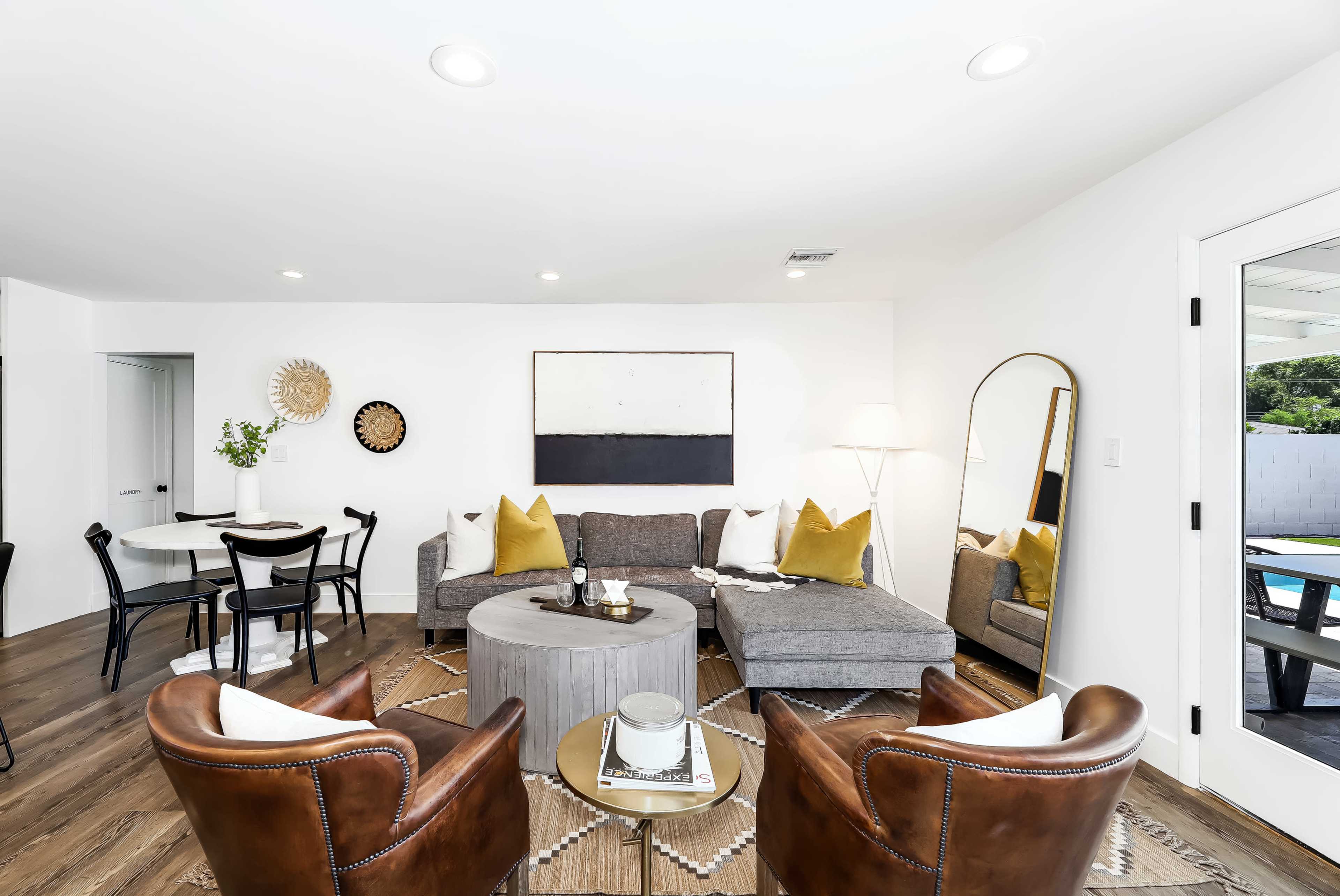 A modern living room featuring a gray sectional sofa, brown leather armchairs, a round coffee table, and a wall art piece, with a dining area visible in the background.