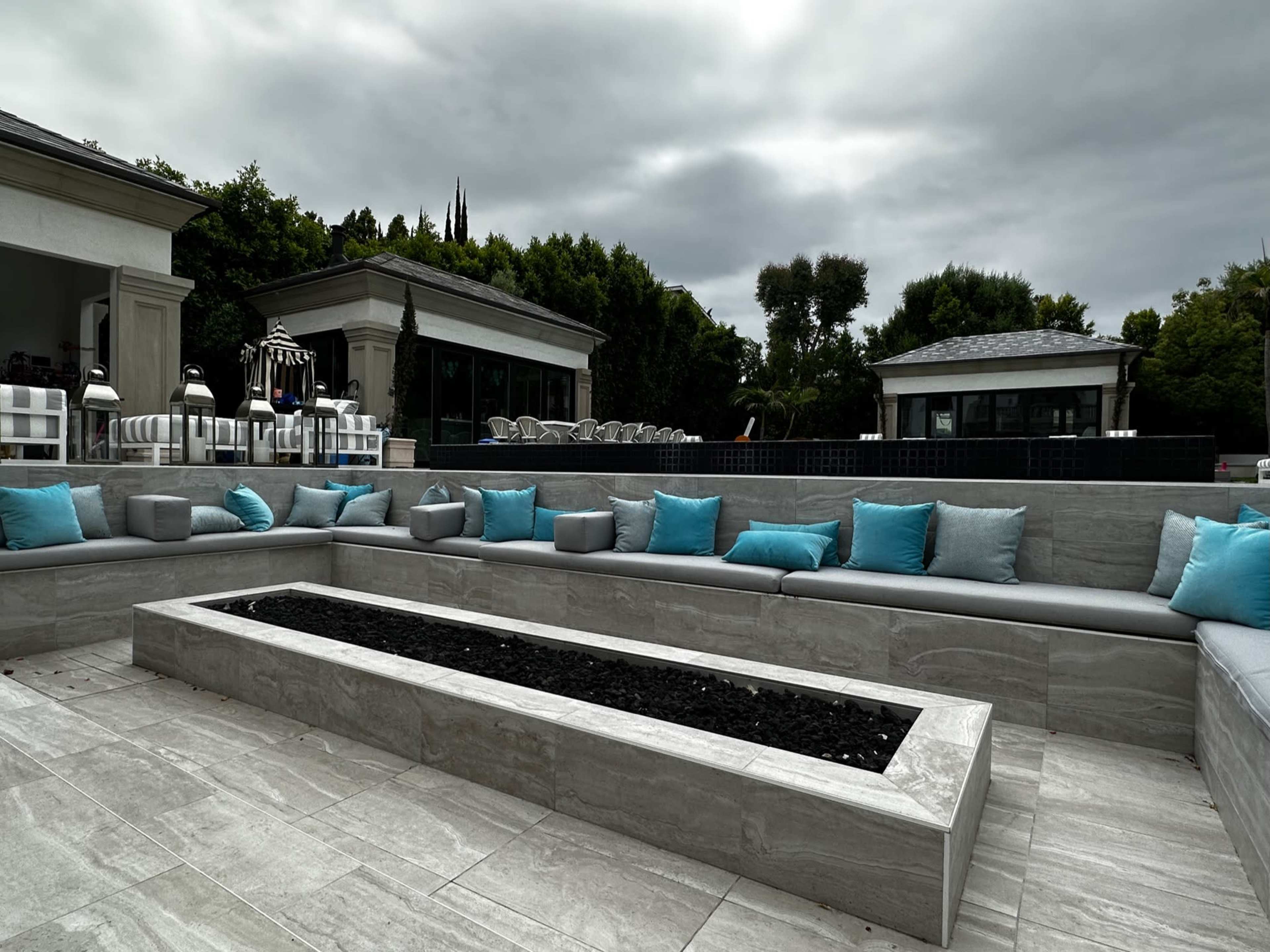 A modern outdoor seating area features gray stone benches with turquoise accent pillows surrounding a low stone firepit.