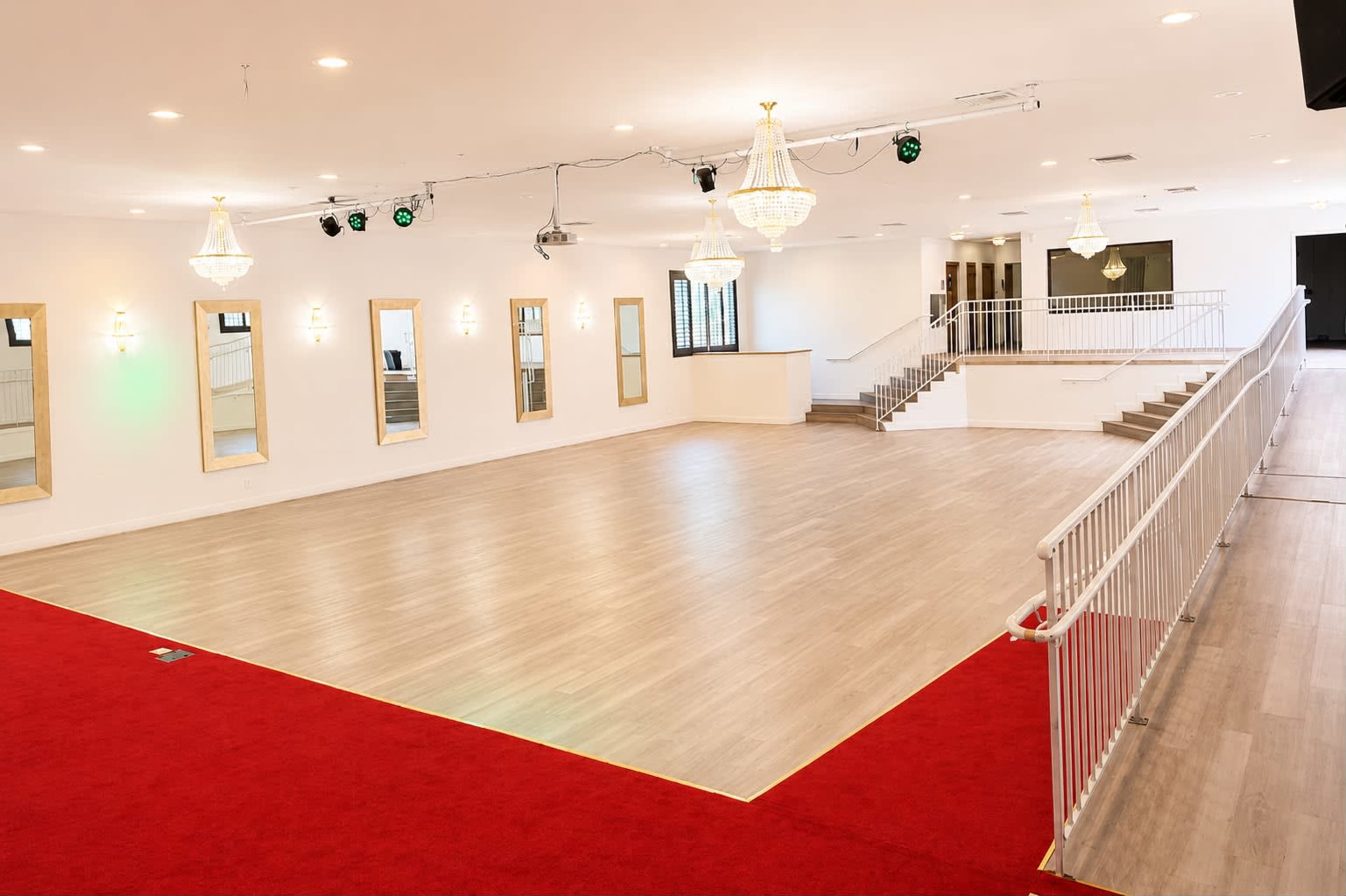 The image shows a spacious event hall with a wooden floor, red carpet, mirrors on the walls, and chandeliers hanging from the ceiling.