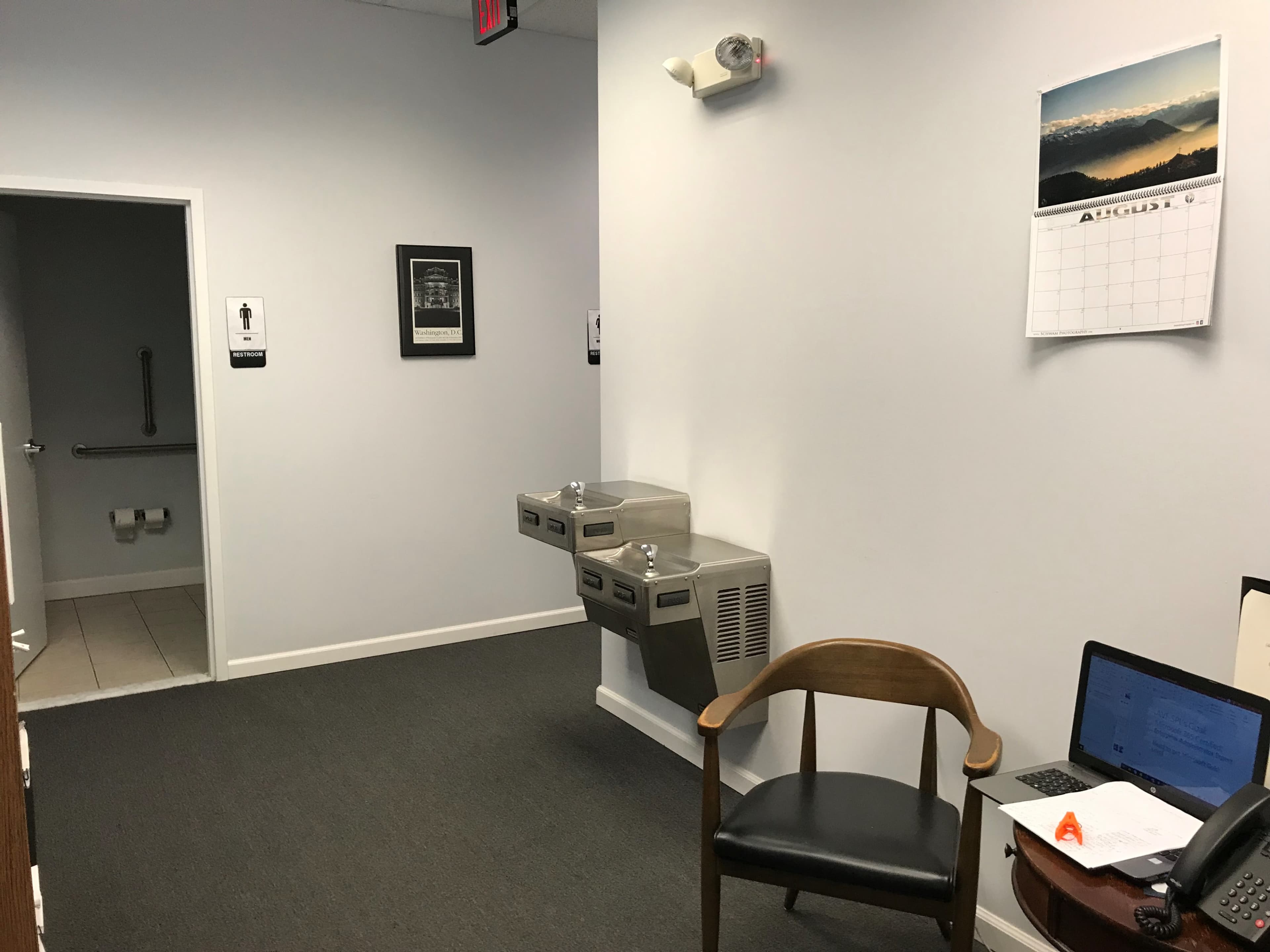 A corridor with a calendar on the wall, two water fountains, a chair, and an open door leading to a restroom.