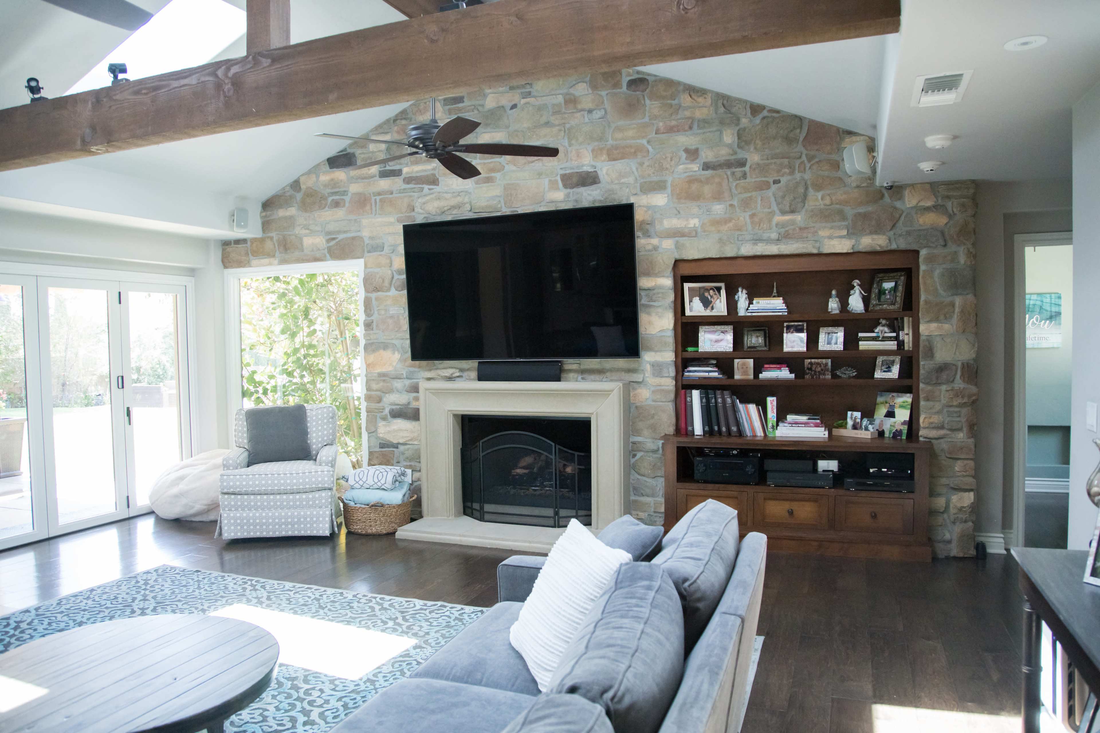 A living room with a stone wall featuring a large TV mounted above a fireplace, flanked by a wooden shelving unit and a cozy seating area.