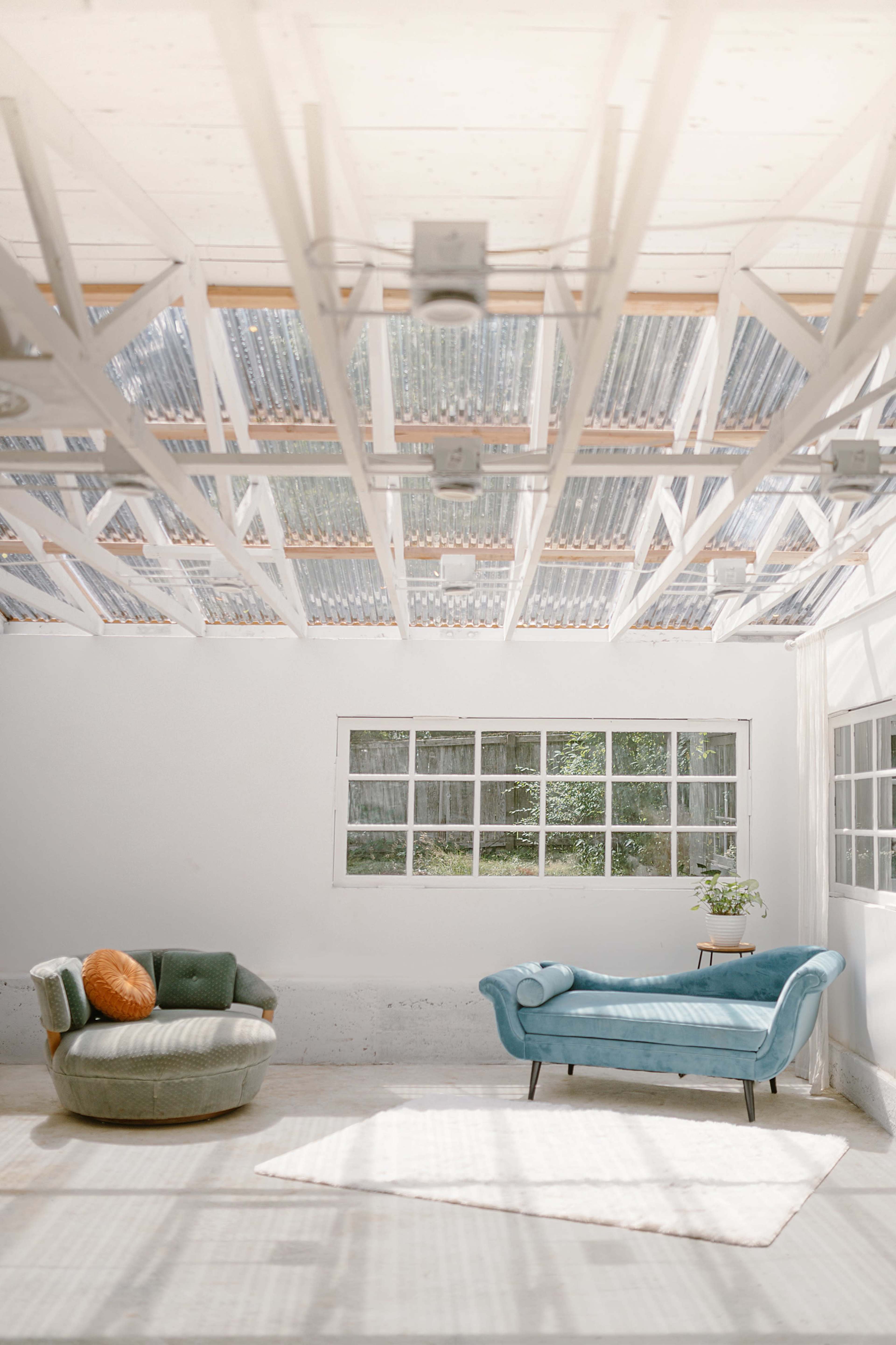 The image shows a light-filled room with a white ceiling and wooden beams, featuring two distinct sofas and a small plant by the window.