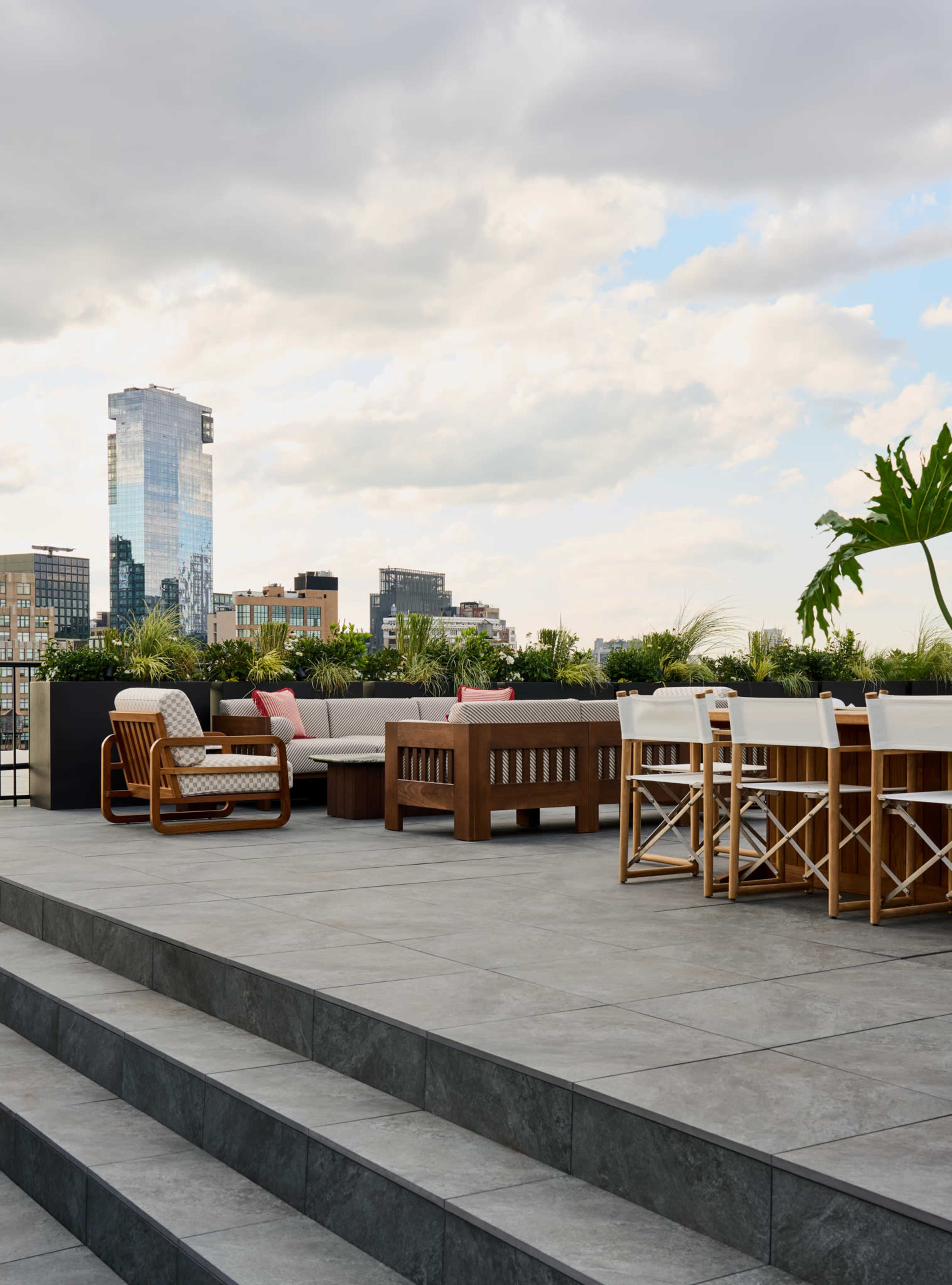 A spacious rooftop terrace features modern furniture, including a wooden dining table and lounge seating, set against a backdrop of city buildings and a cloudy sky.