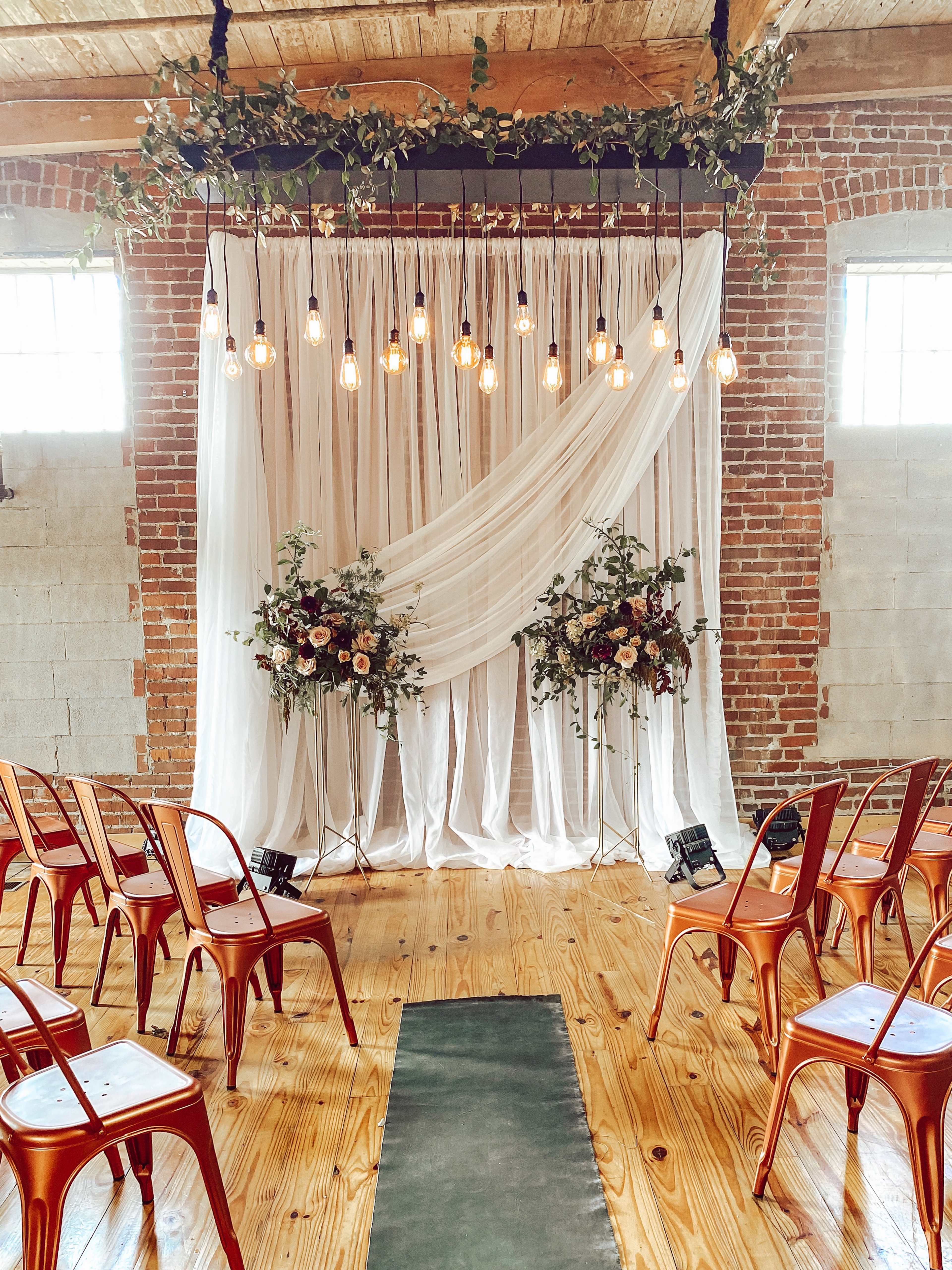 A wedding ceremony setup with a decorative fabric backdrop, hanging lights, floral arrangements, and rows of chairs arranged on a wooden floor.