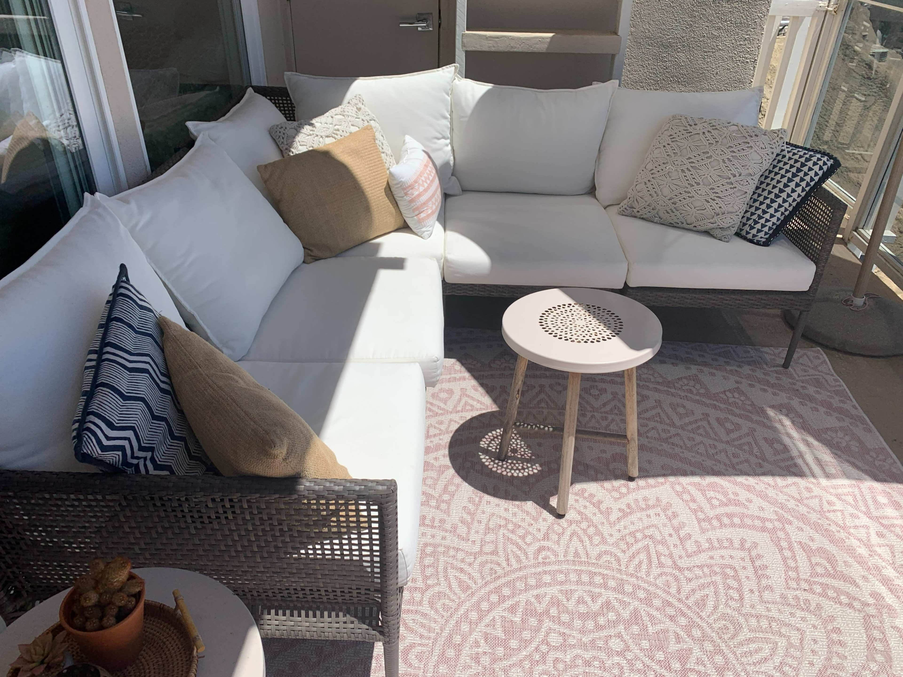 A cozy outdoor seating area featuring a sectional couch with white cushions, decorative pillows, and a small round table on a patterned rug.