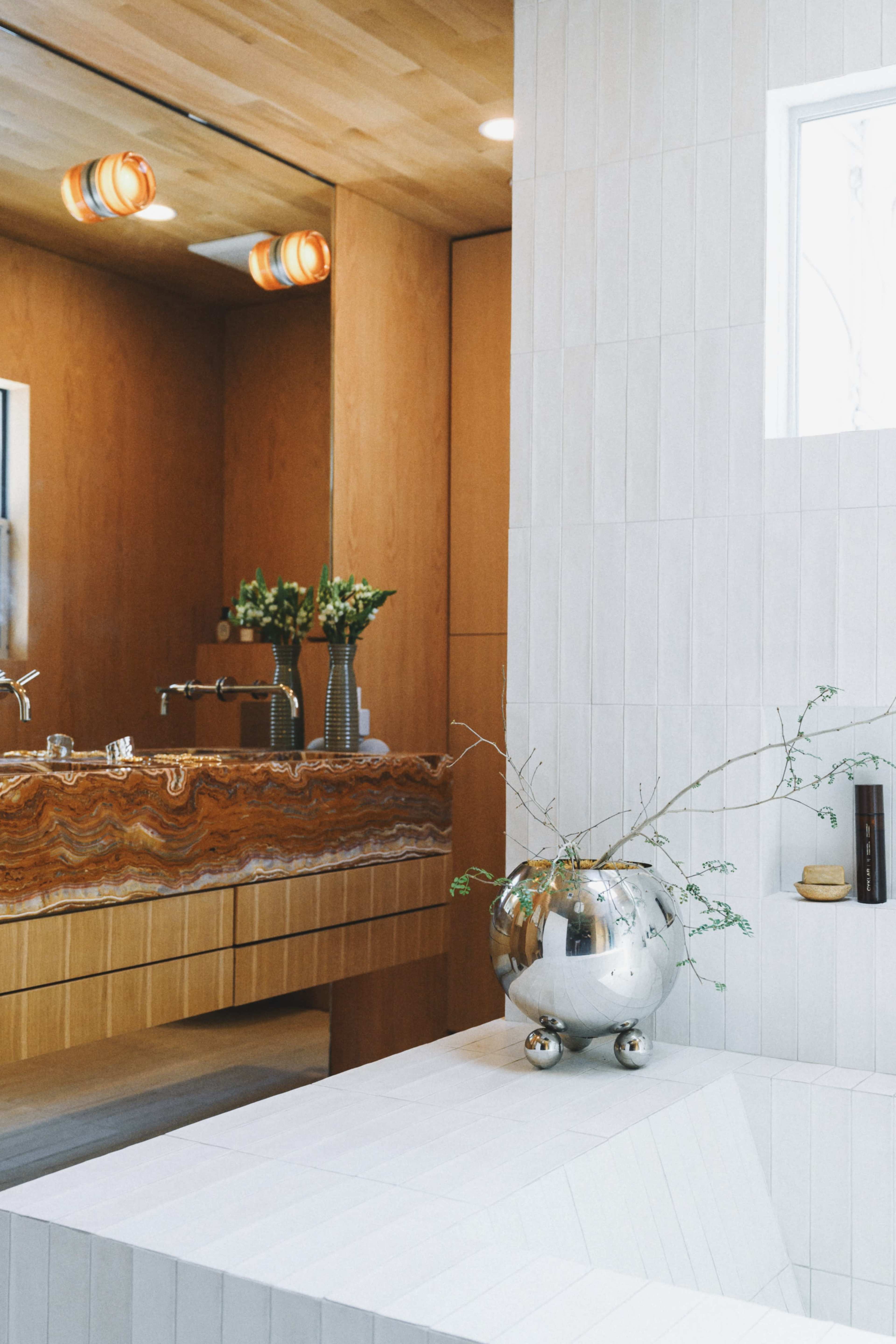 A modern bathroom features a mirrored vanity with a wooden cabinet, a striking onyx countertop, and a minimalist bathtub adorned with a round silver planter.
