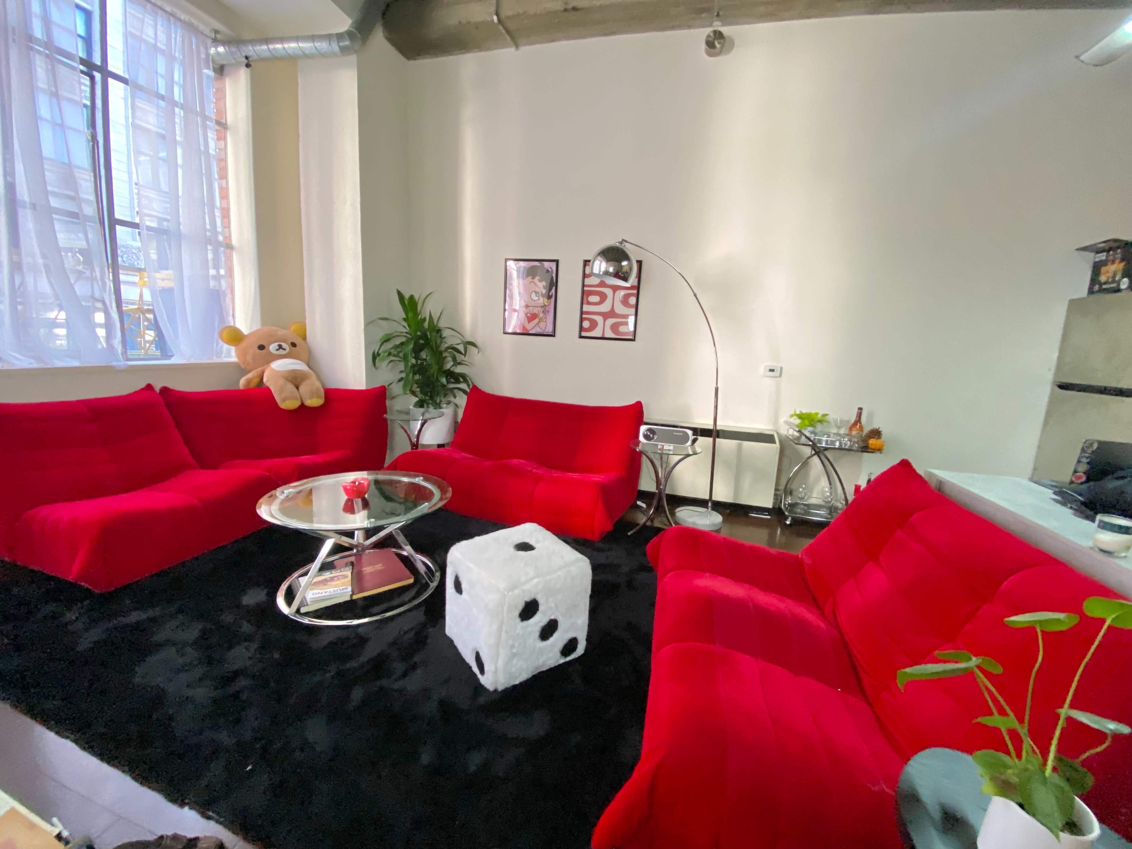 The room features red sectional sofas arranged around a round glass coffee table, with a white plush cube and a teddy bear on one sofa.