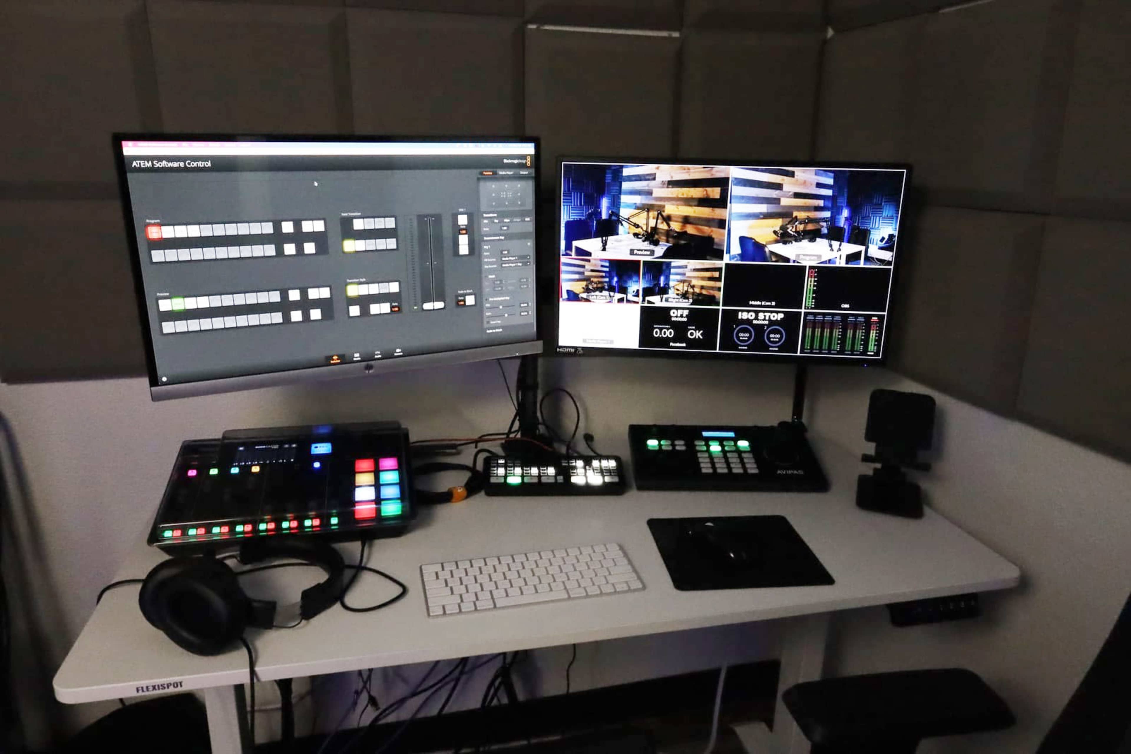 A dual-monitor setup on a desk with broadcast control software displayed, along with a control panel and headphones.