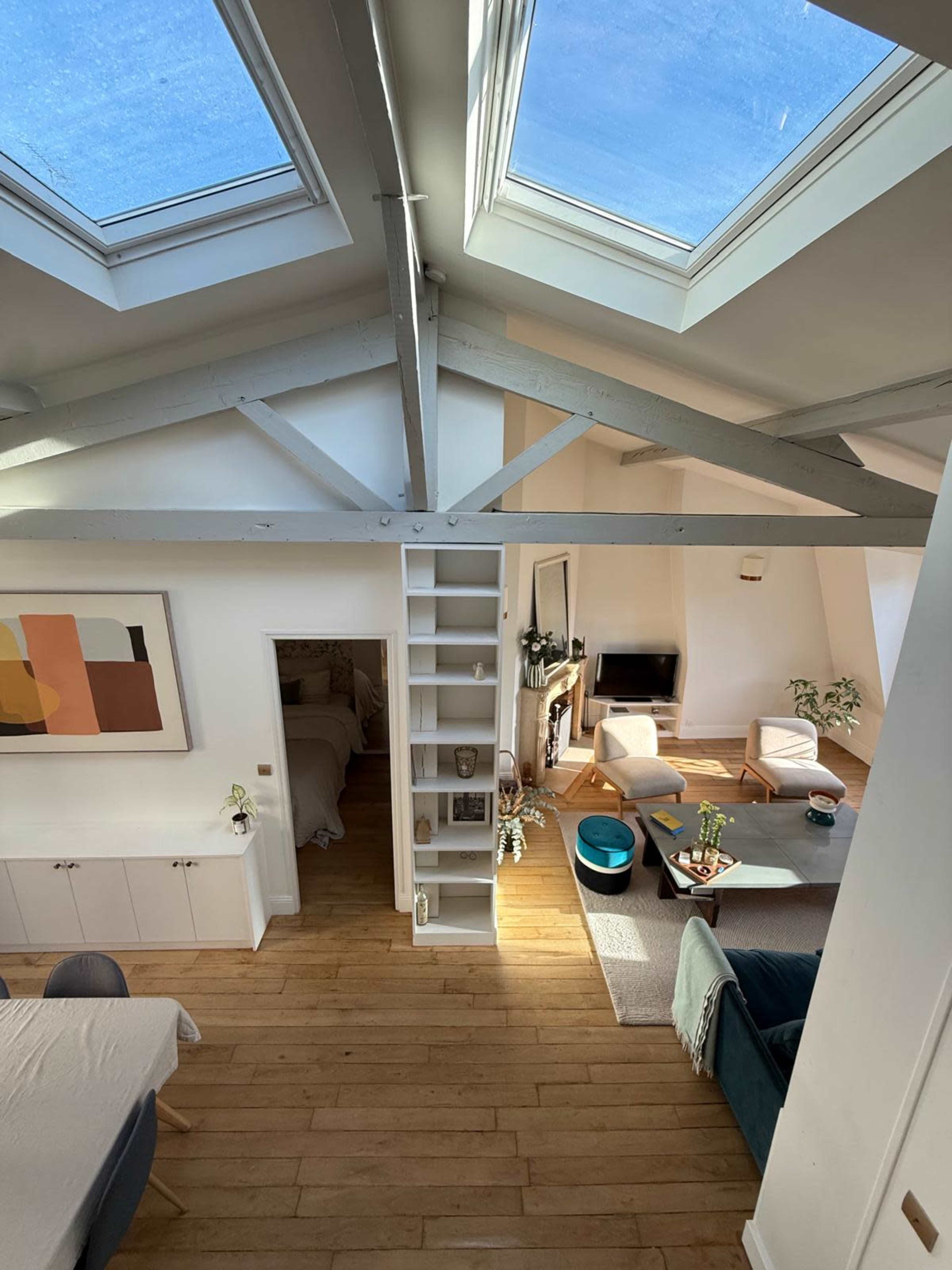 The image shows a spacious loft-style living area with large skylights, wooden beams, and a mix of furniture including a sofa and a dining table.