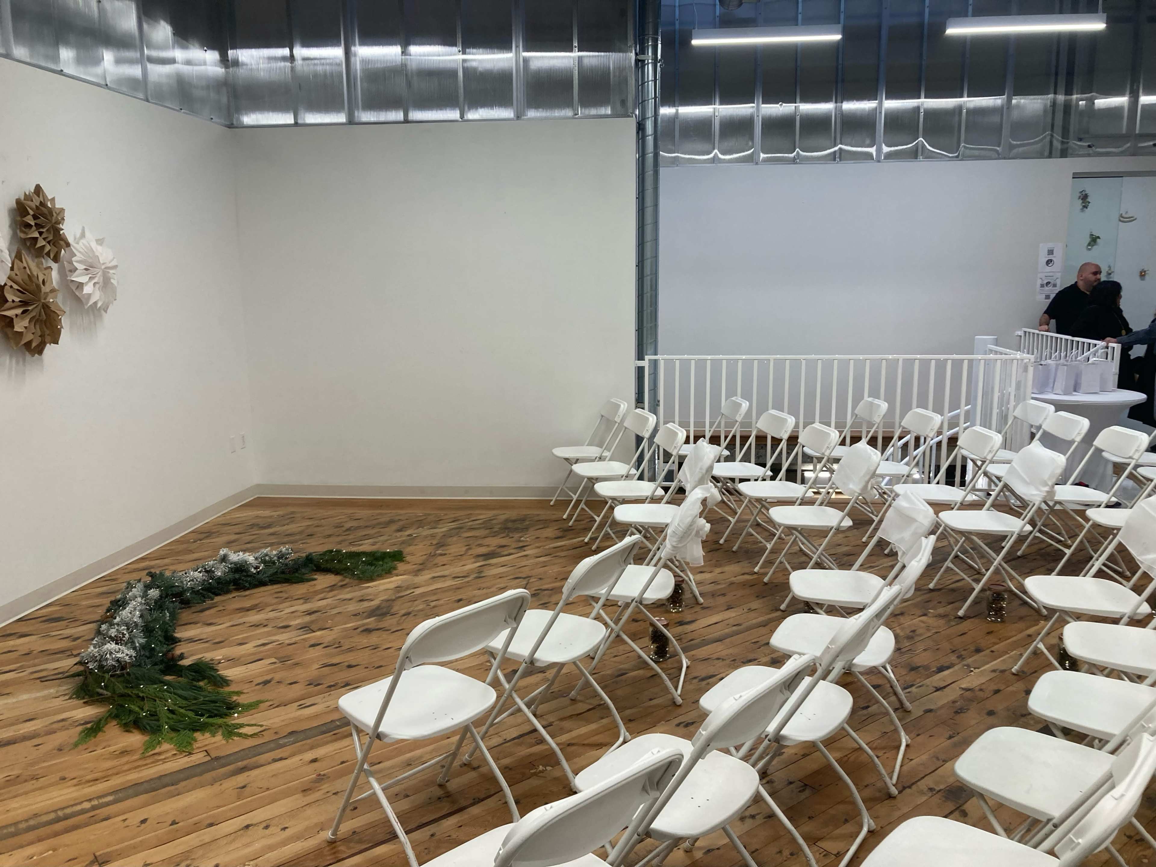A room features white folding chairs arranged in rows facing a decorative arrangement of greenery and paper crafts on the wooden floor.