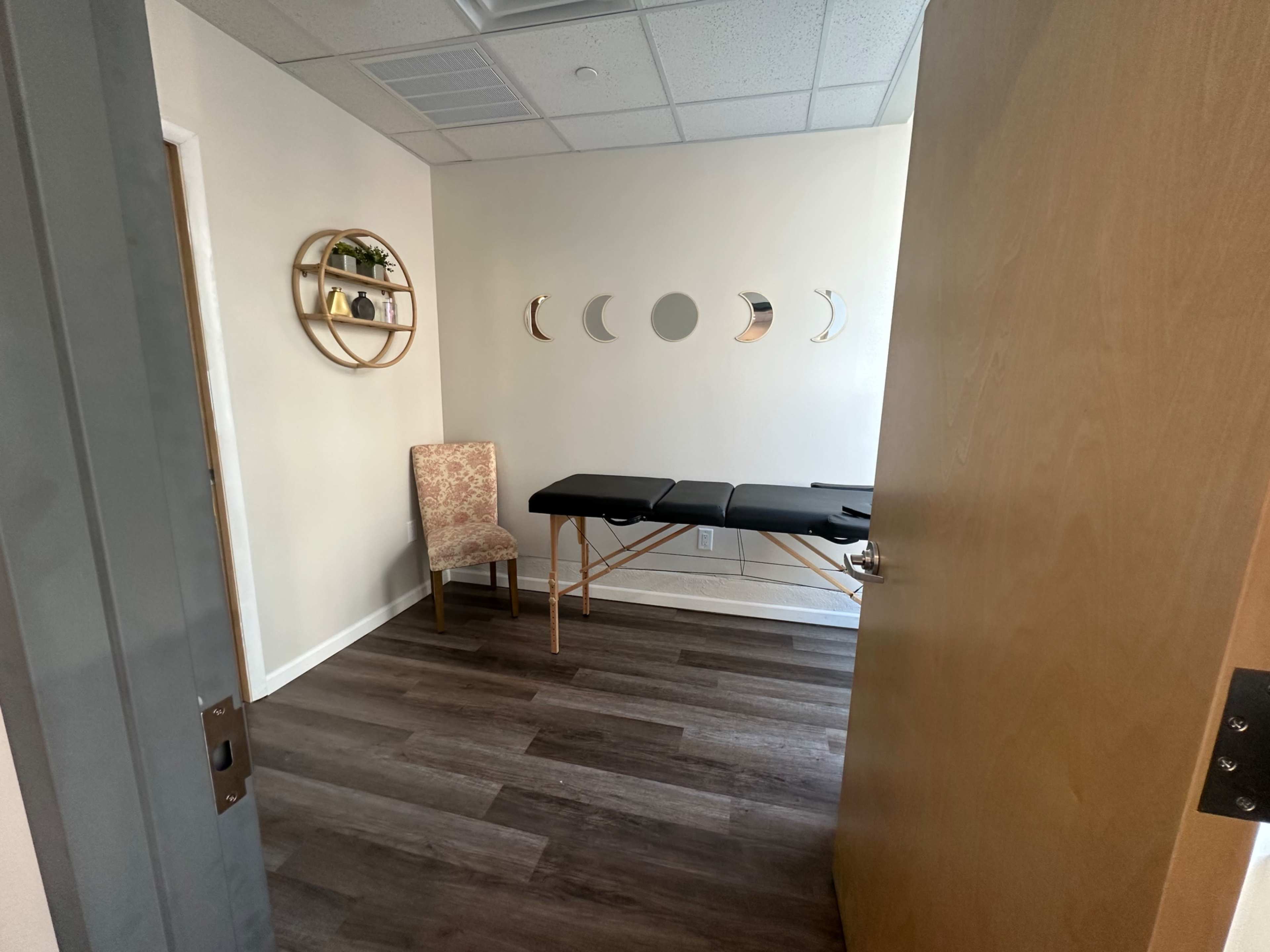 A small treatment room with a massage table, a chair, and a decorative wall featuring circular mirrors and plants.
