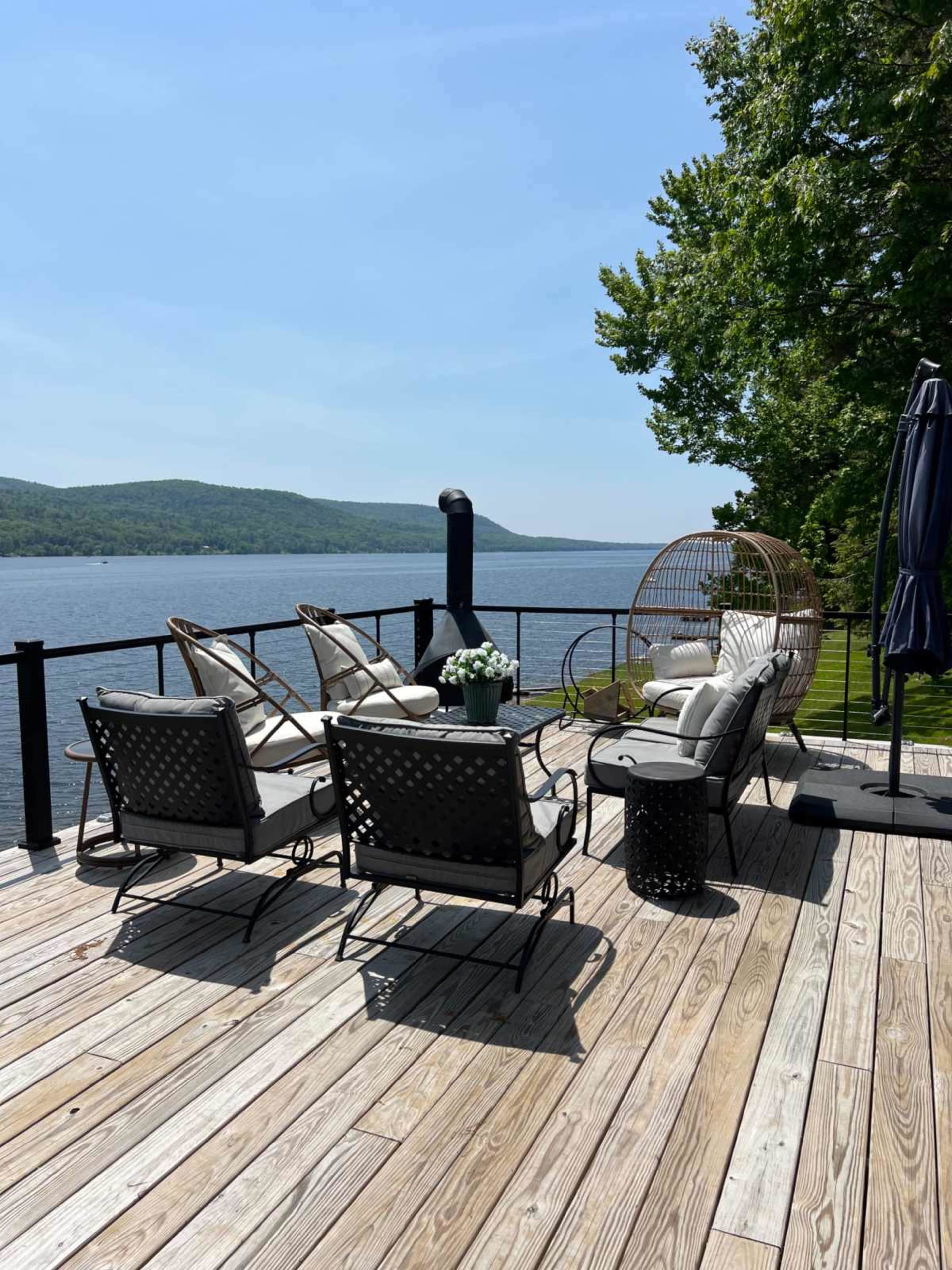 A wooden deck overlooks a calm lake, featuring several lounge chairs, a round table with flowers, and an outdoor umbrella.
