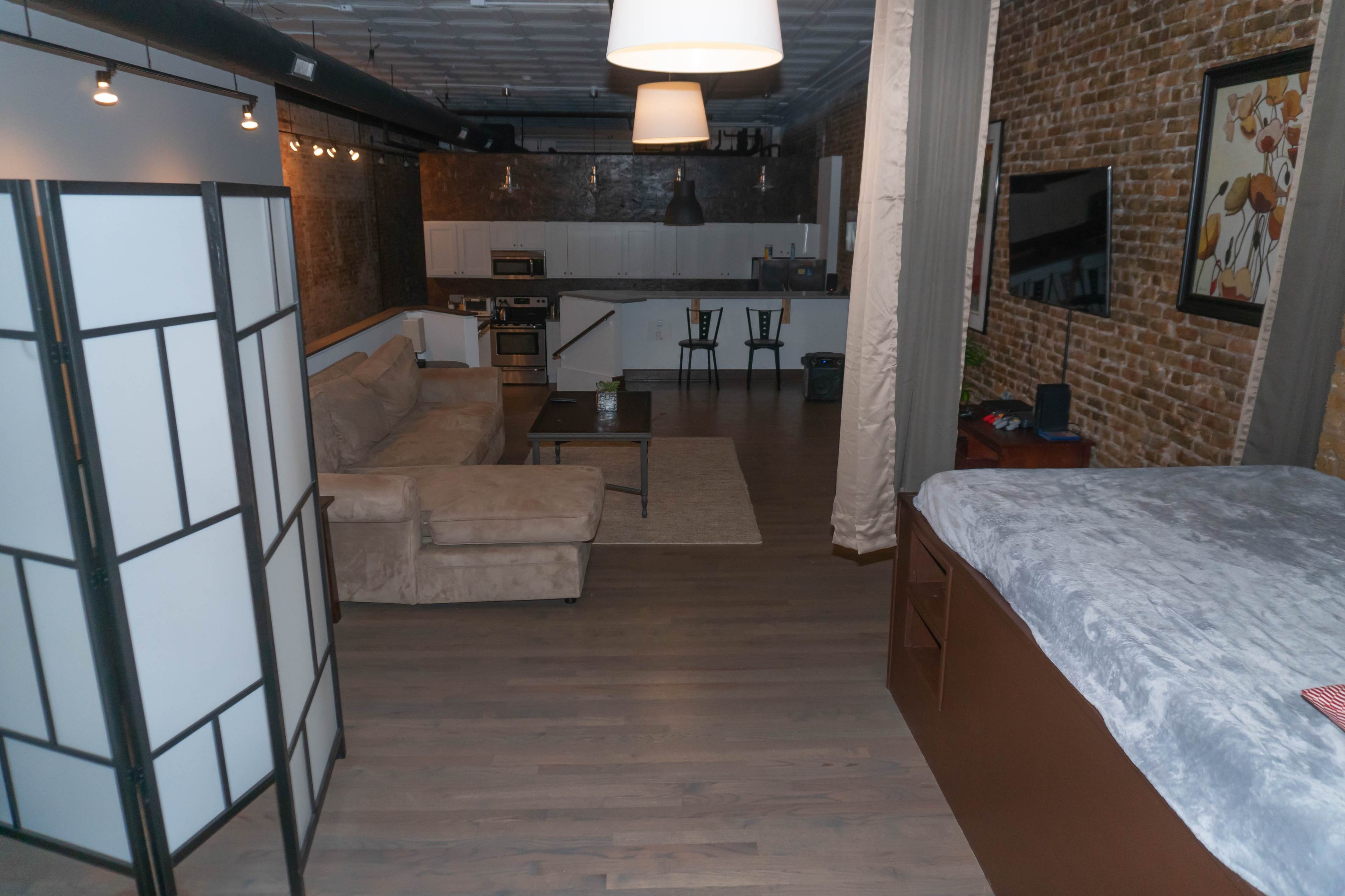 An open loft-style living space featuring a living area with a couch, a kitchenette in the background, and a bed area with a grey blanket.