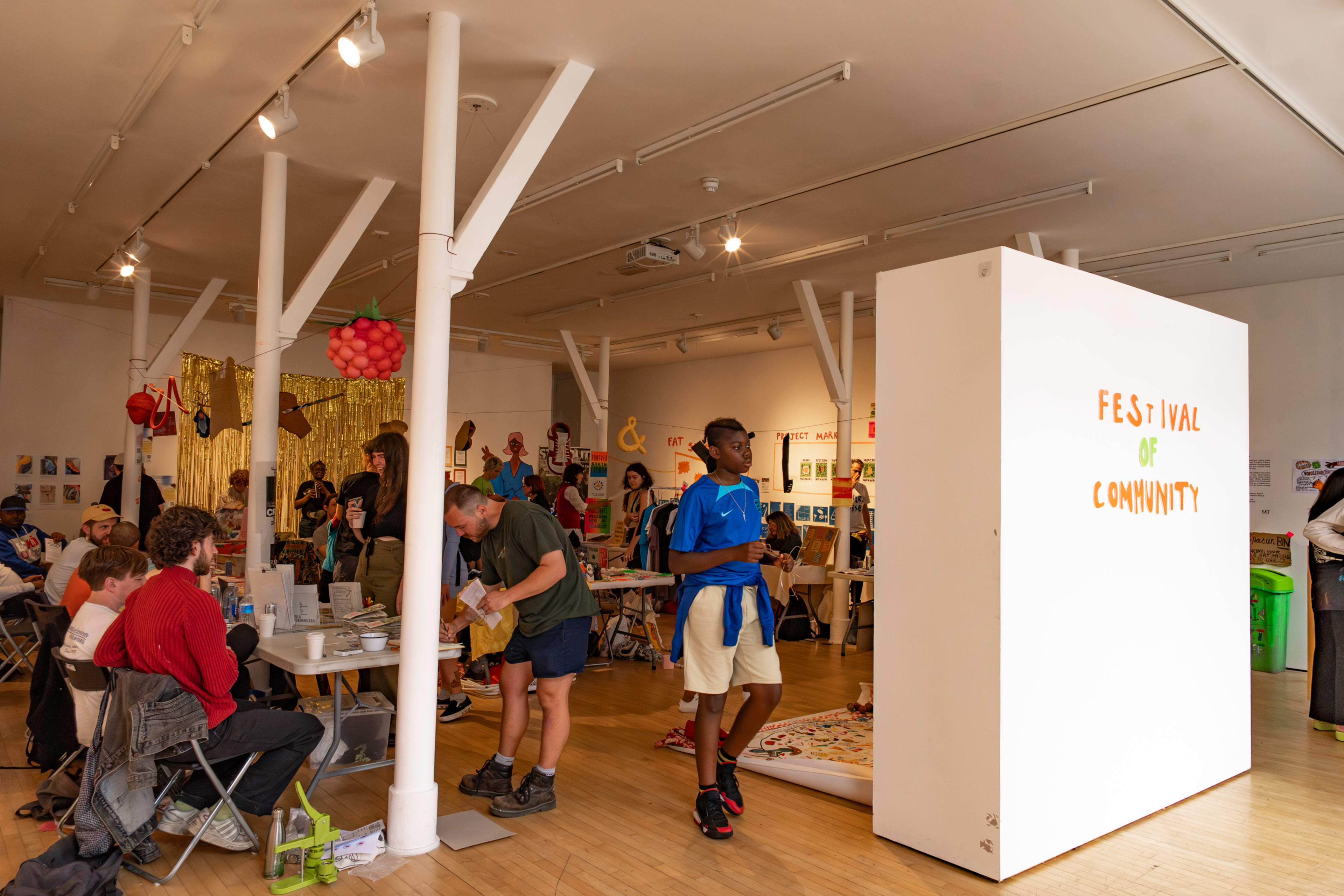 The image shows a community event space filled with people engaging in activities, with a prominent white wall displaying the words "FESTIVAL OF COMMUNITY."