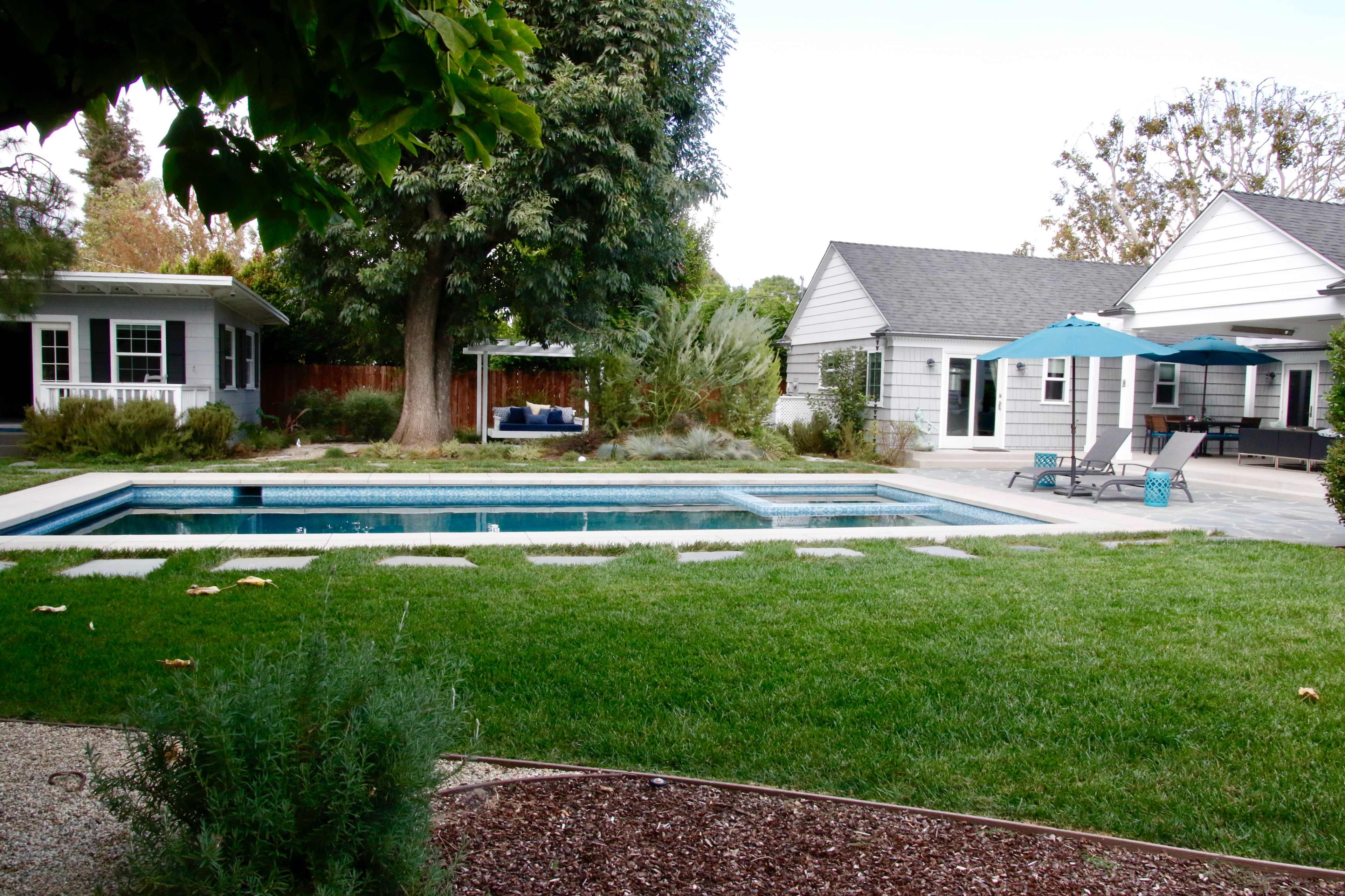 A neatly landscaped backyard features a swimming pool, bordered by grassy areas and stone pathways, with a patio and a nearby shaded seating area.