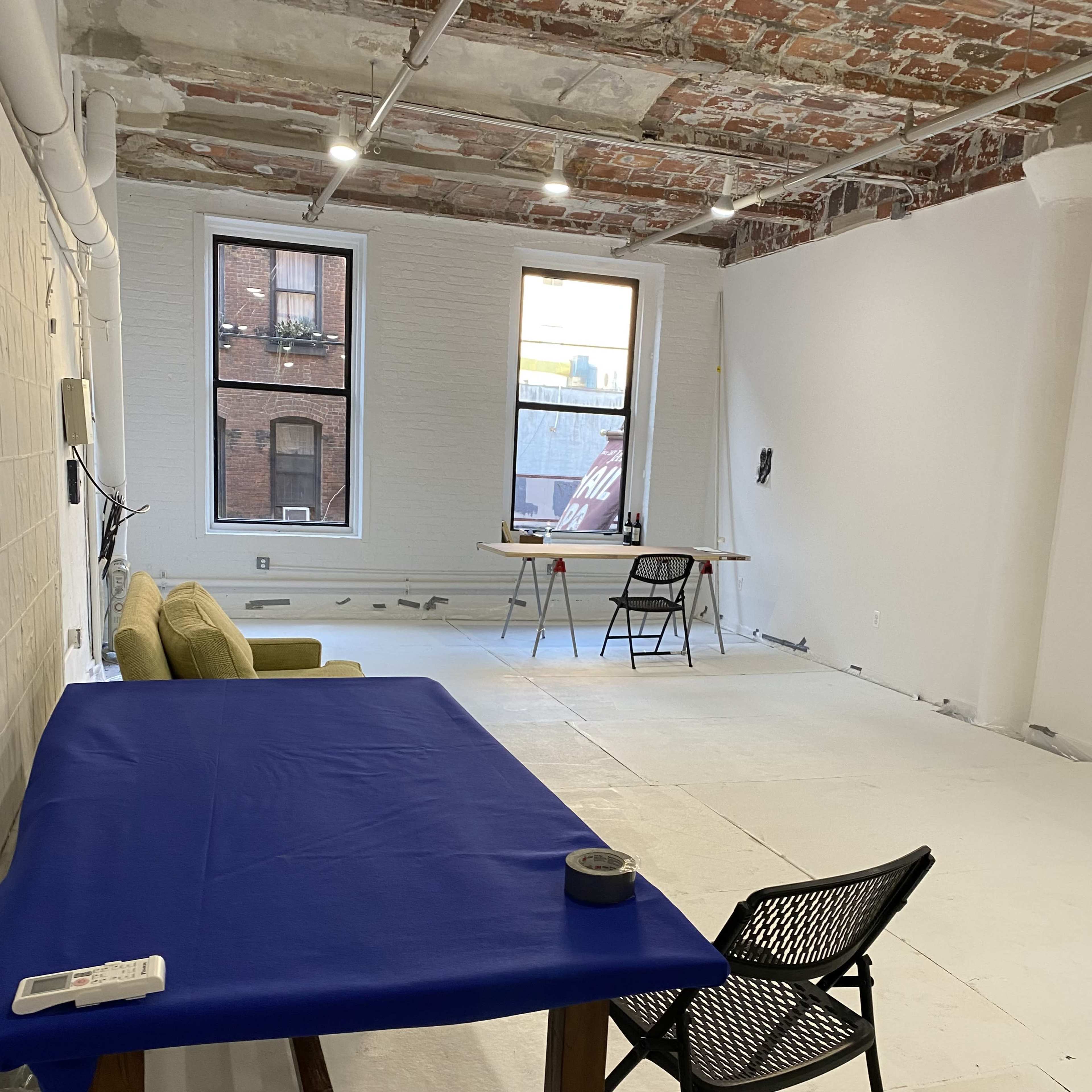 An unfinished room with exposed brick walls, a blue table, a chair, and a small work area with tools near a window.