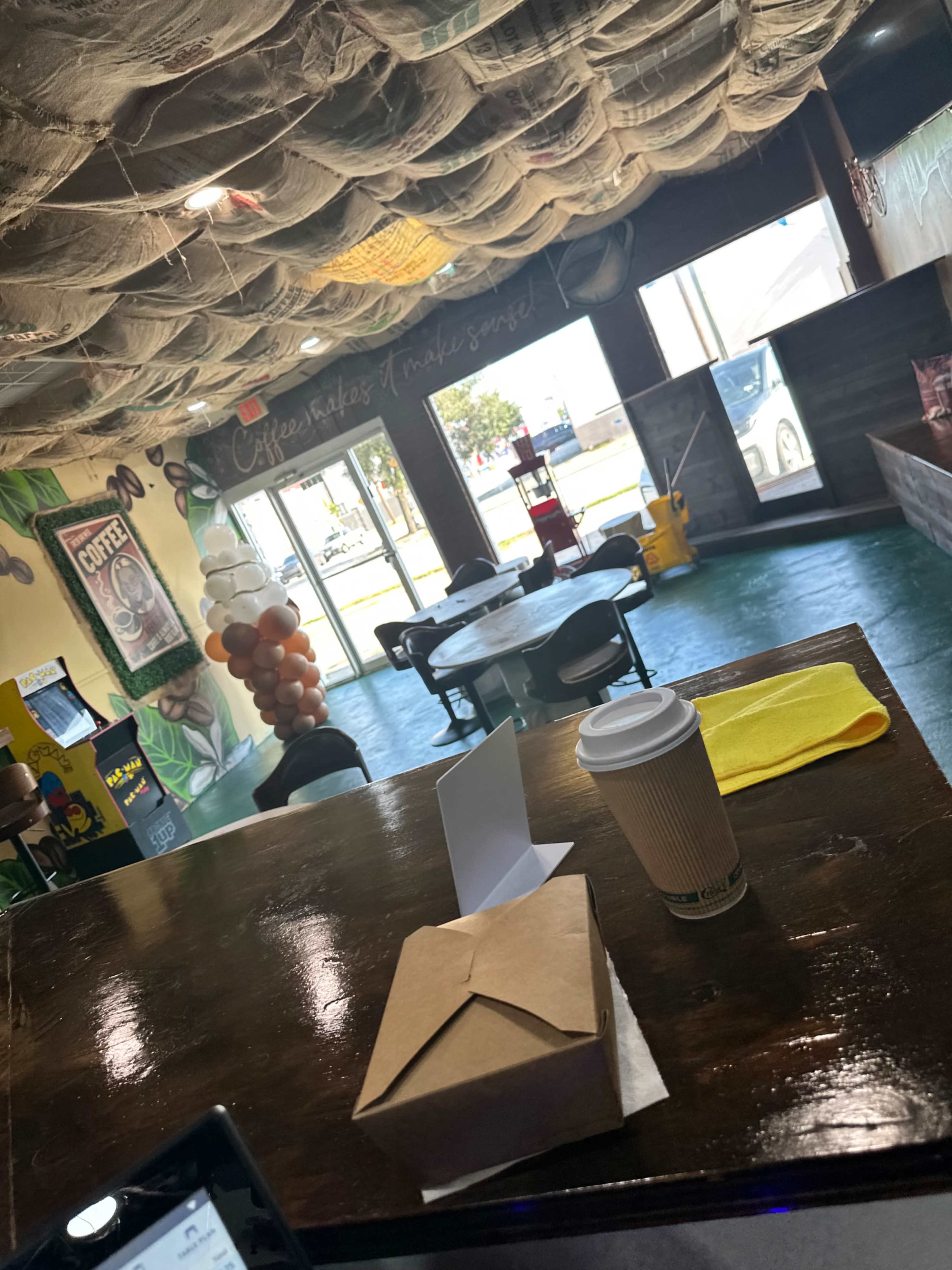 The image shows the interior of a coffee shop with a wooden table foreground, a coffee cup, and takeout box, while the decor includes a ceiling covered in netting and colorful balloons near the entrance.