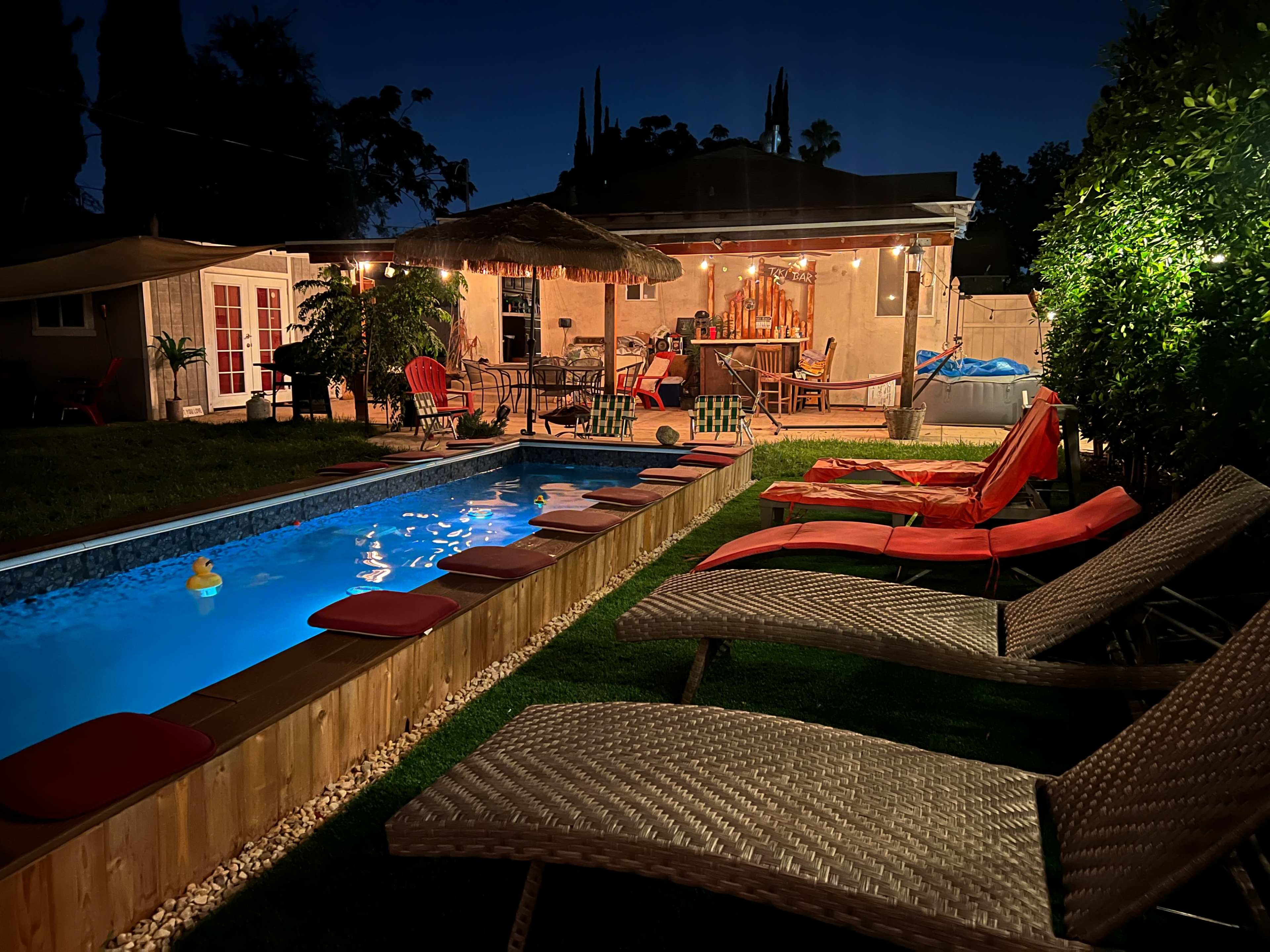 A well-lit backyard pool area at night, with lounge chairs lining the pool and a patio with seating and decorative lights in the background.
