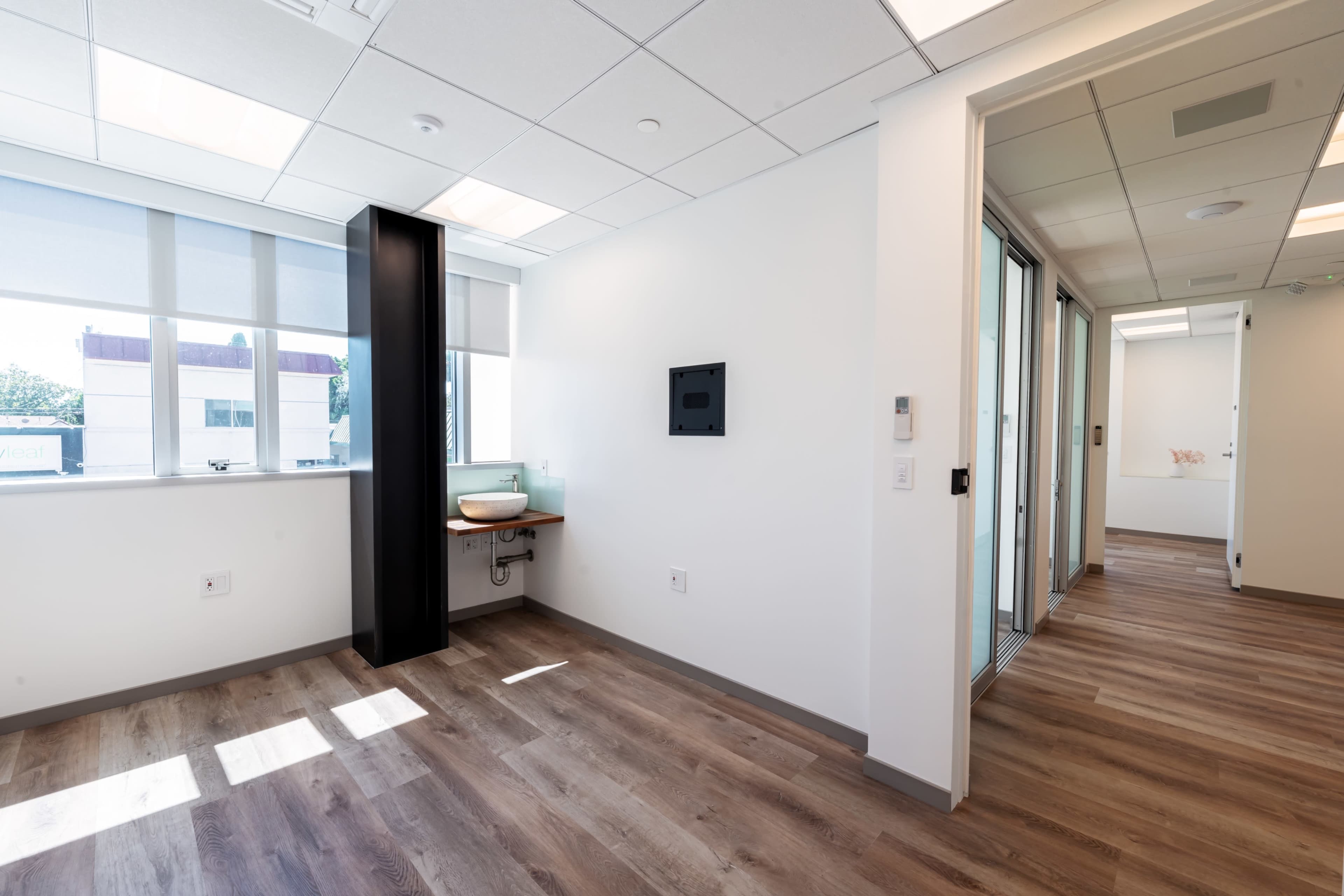 A brightly lit office space with large windows, wooden flooring, and minimal furnishings including a wall-mounted screen.