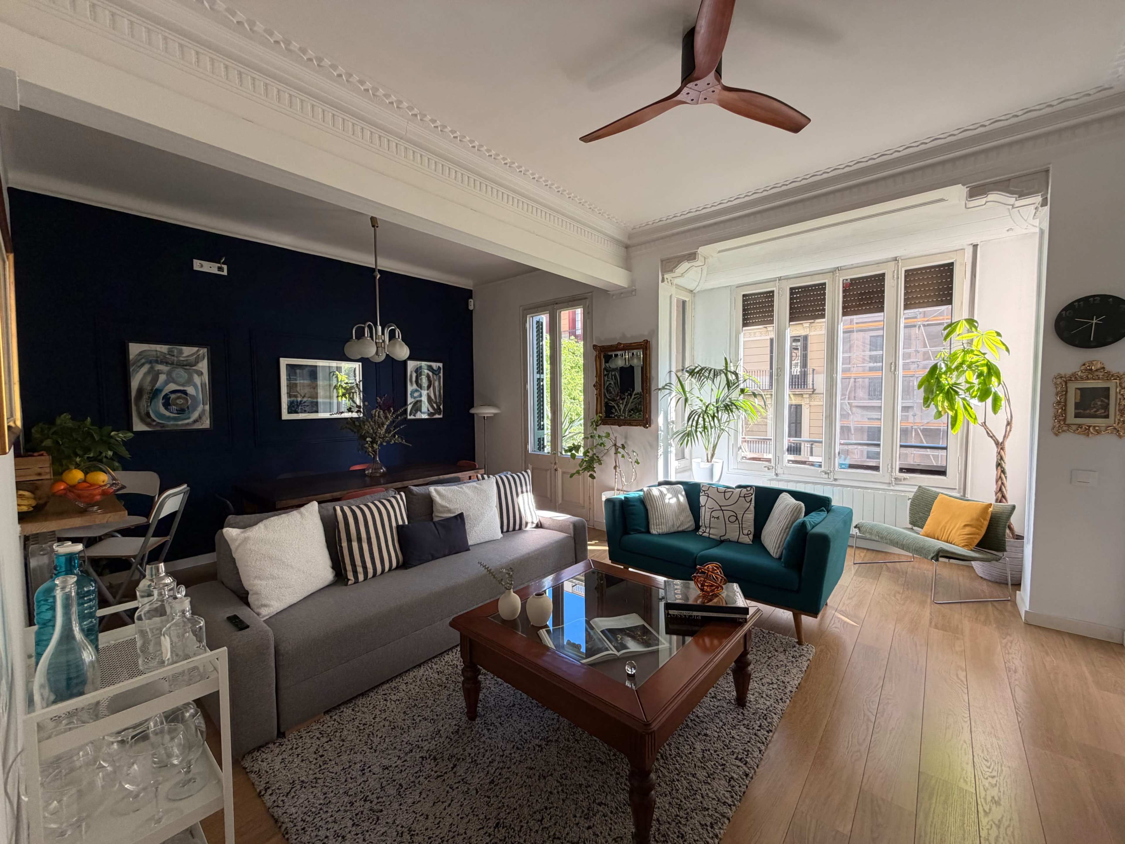 The image shows a modern living room featuring a gray sofa, a turquoise armchair, a wooden coffee table, and large windows with natural light illuminating the space.
