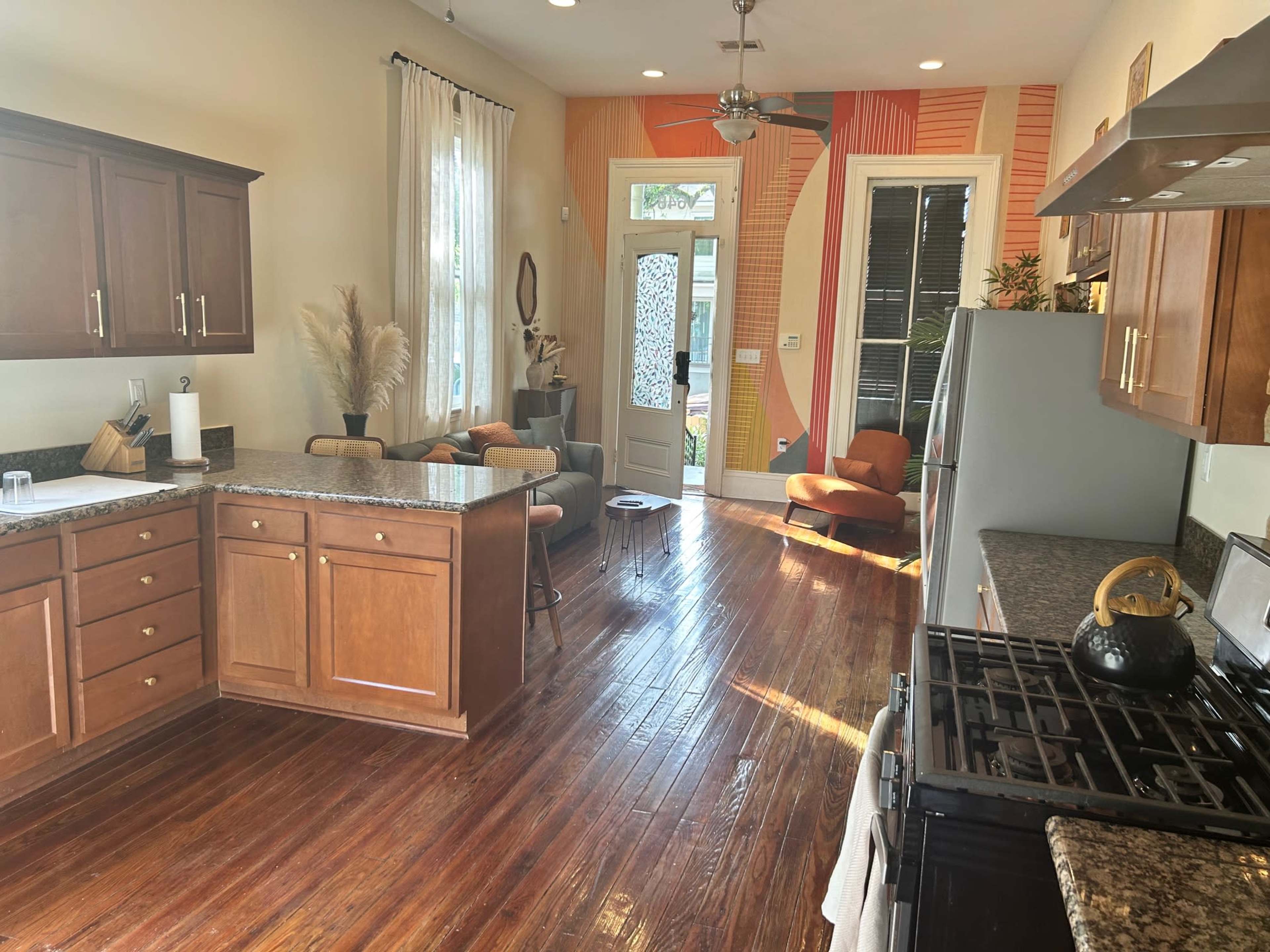 A modern kitchen with wooden cabinets, a granite countertop, and a view into a brightly decorated living space through a glass door.