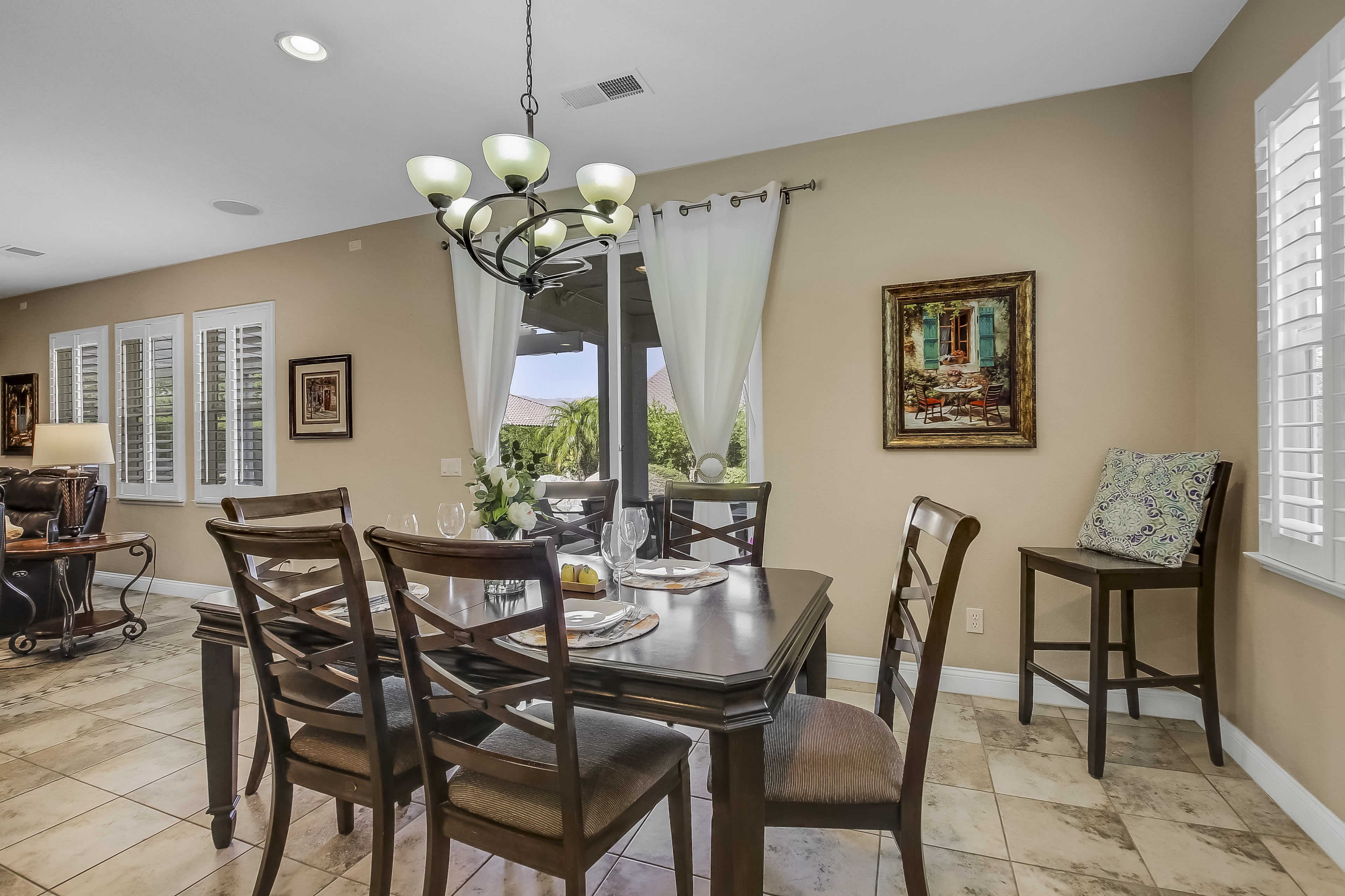 A dining area features a wooden table set for a meal, surrounded by chairs, with a chandelier overhead and windows letting in natural light.