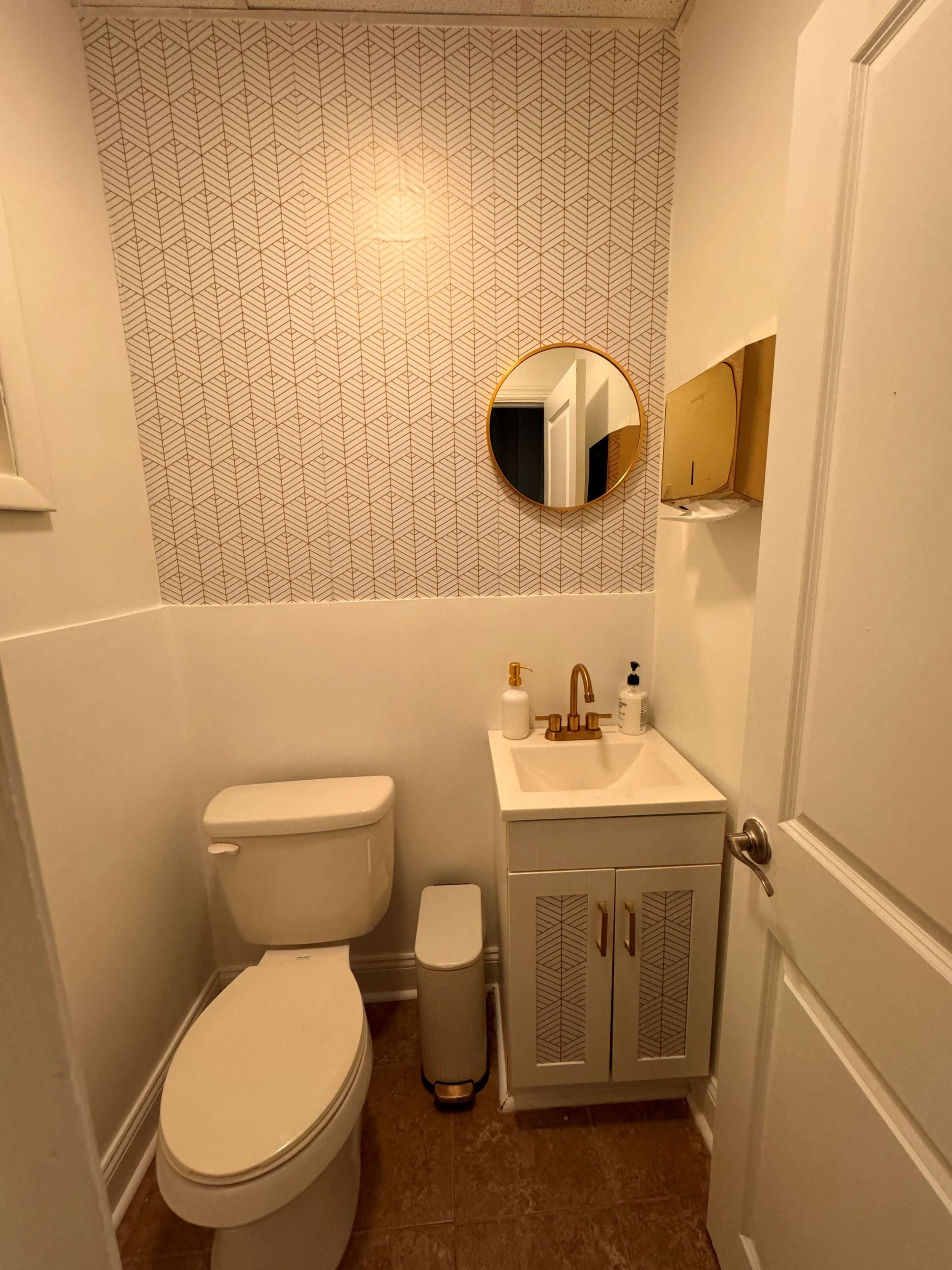 A small bathroom features a toilet, a sink with a cabinet, and a round mirror above the sink, all set against a patterned wallpaper.
