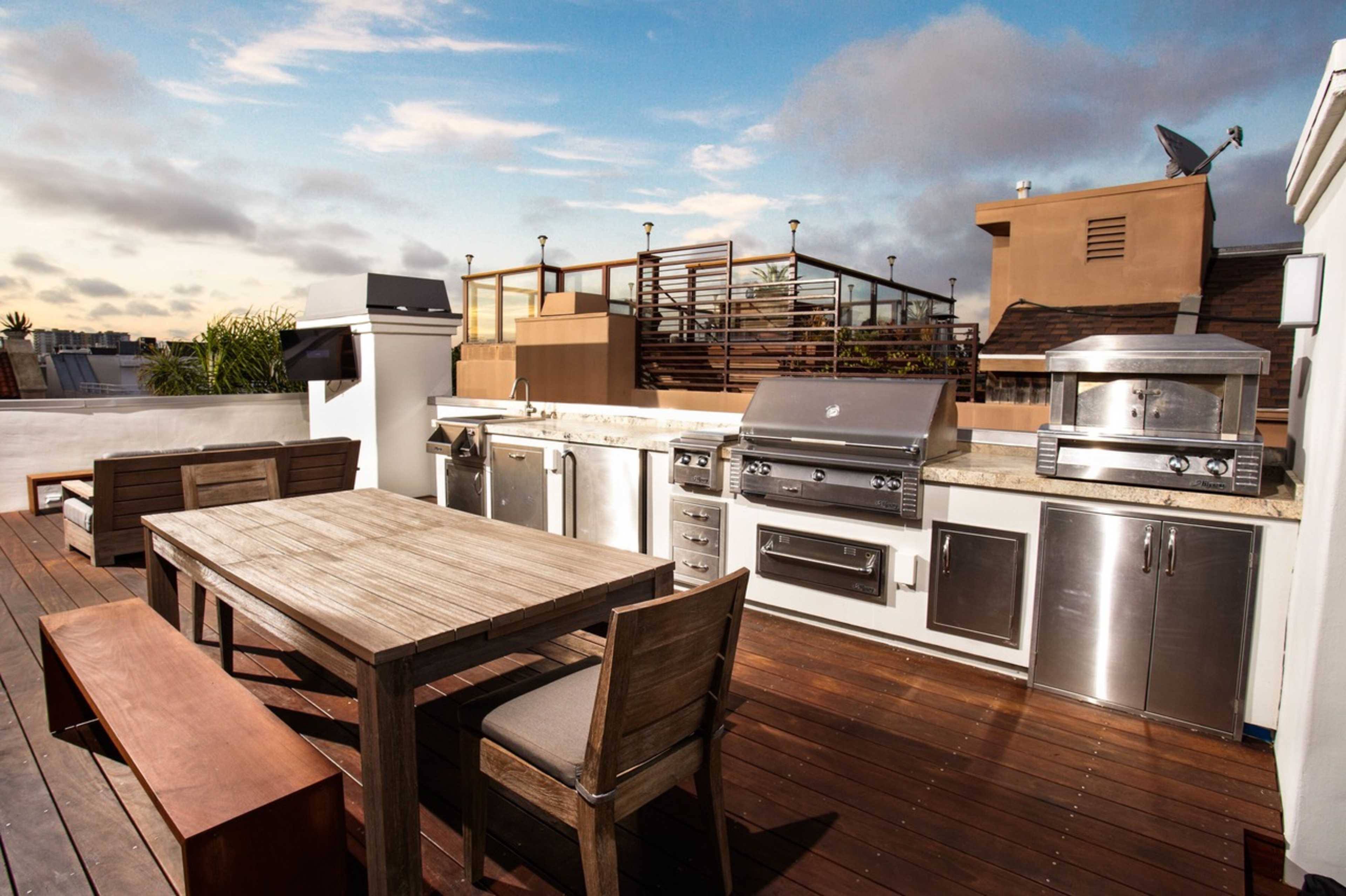 The image shows a rooftop patio with a barbecue grill, outdoor kitchen appliances, a wooden dining table, and seating areas surrounded by a skyline view.